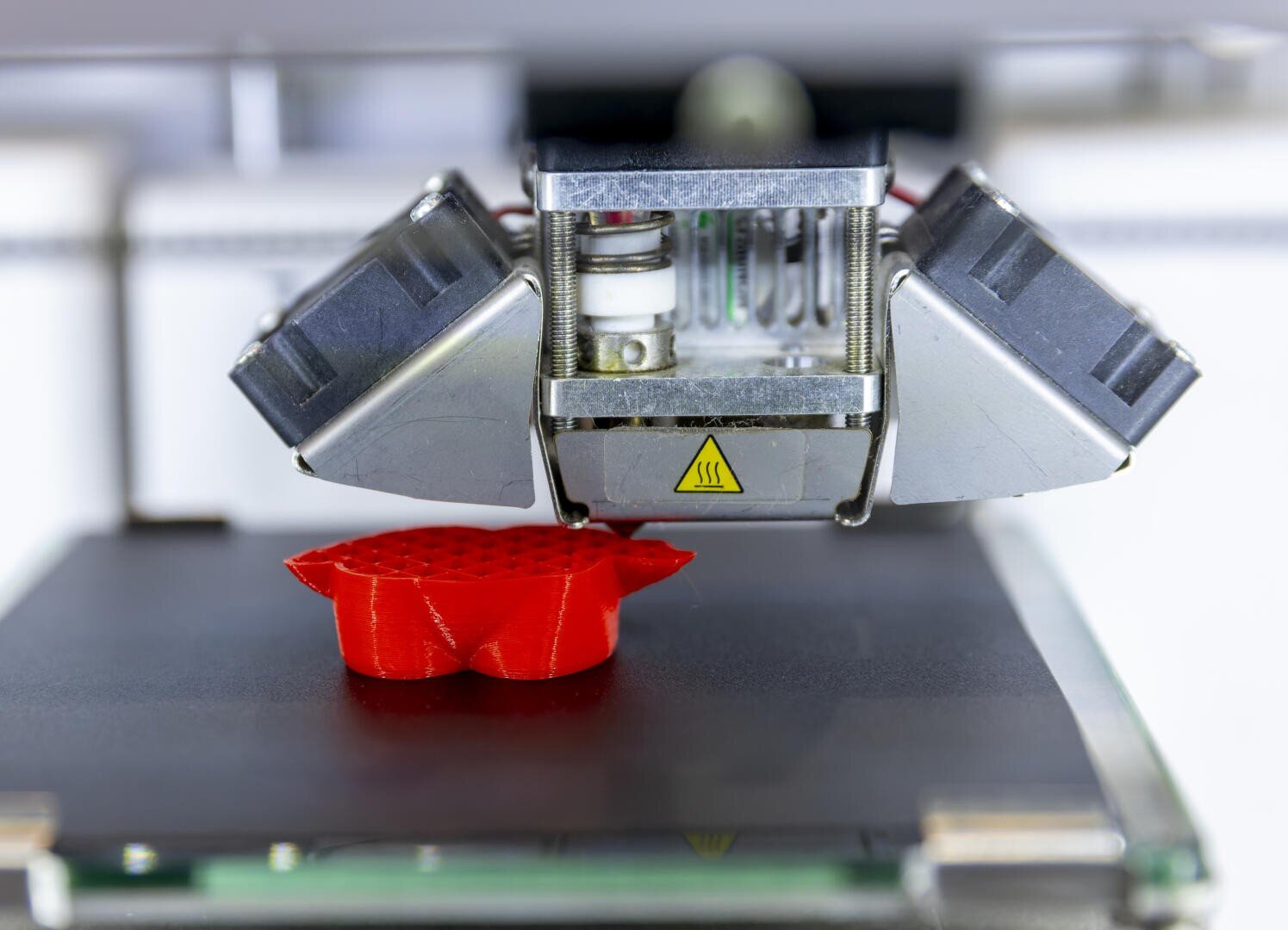 Nahaufnahme einer 3D-Druckerdüse, die ein rotes Objekt in Form einer Blume oder eines Zahnrads auf das Druckbett druckt. Der Druckkopf und das teilweise fertige Objekt sind im Fokus.