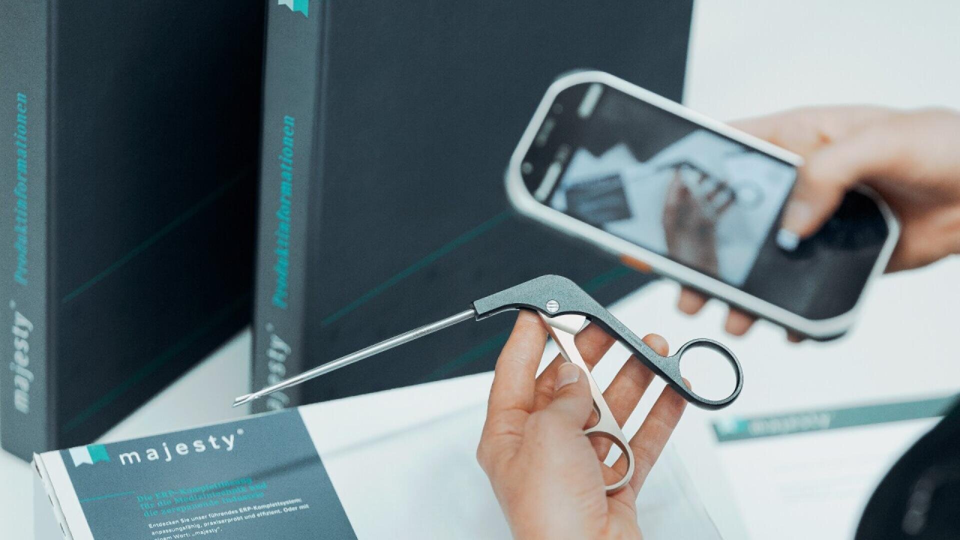 A person holds a pair of scissors over a majesty branded box while taking a photo with a smartphone. More boxes are visible in the background.