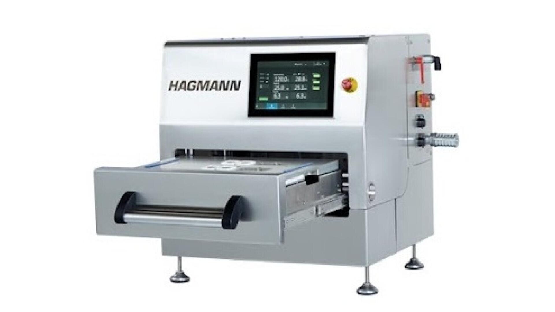 A silver HAGMANN industrial machine with a touchscreen display and an open front drawer, standing on four legs, used for automated manufacturing or processing tasks.