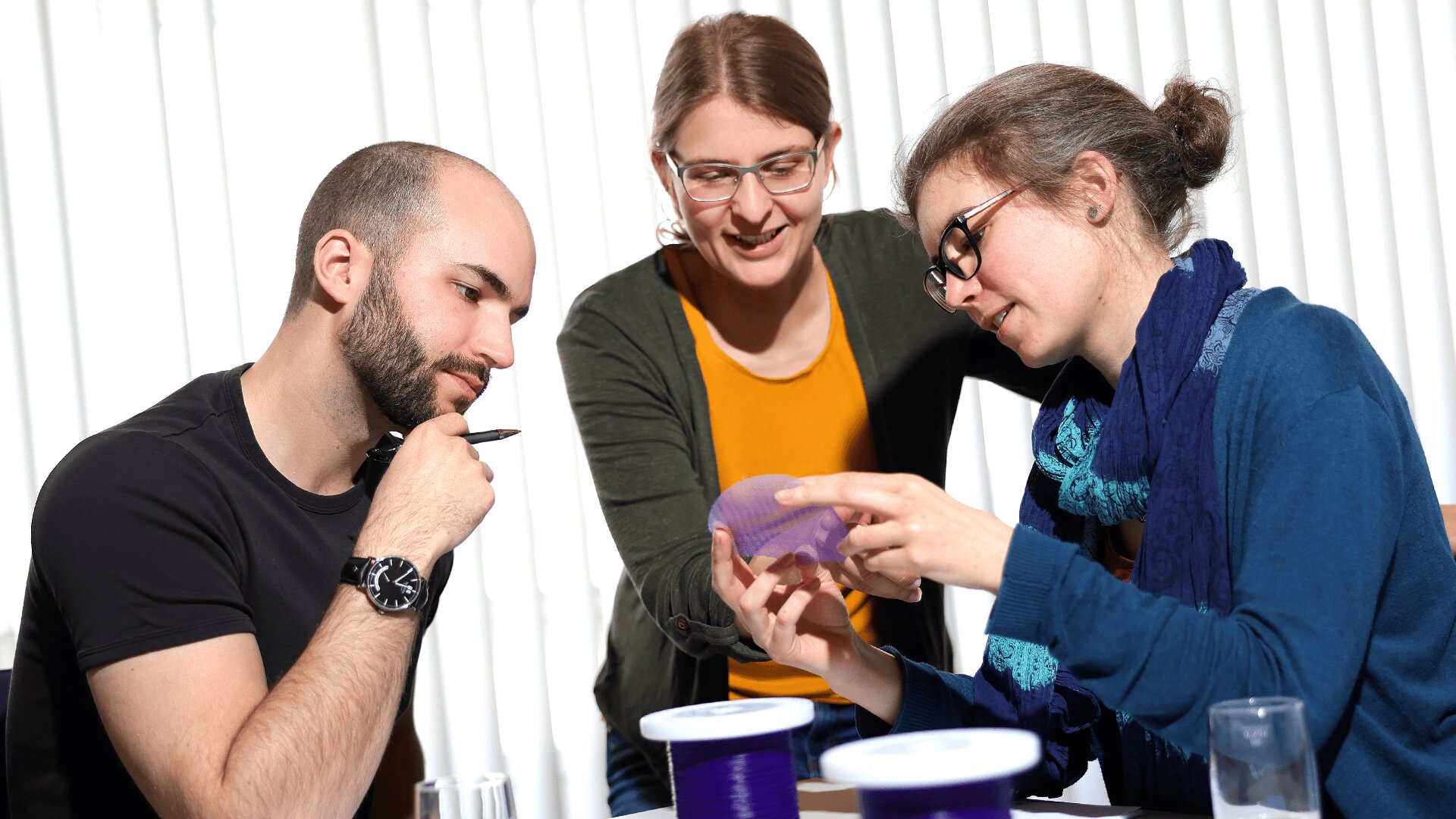 Three people sit at a table examining a translucent purple object. One person holds it while another points, and the third watches thoughtfully with a pen. Spools of filament are visible on the table. Bright light comes from behind them.