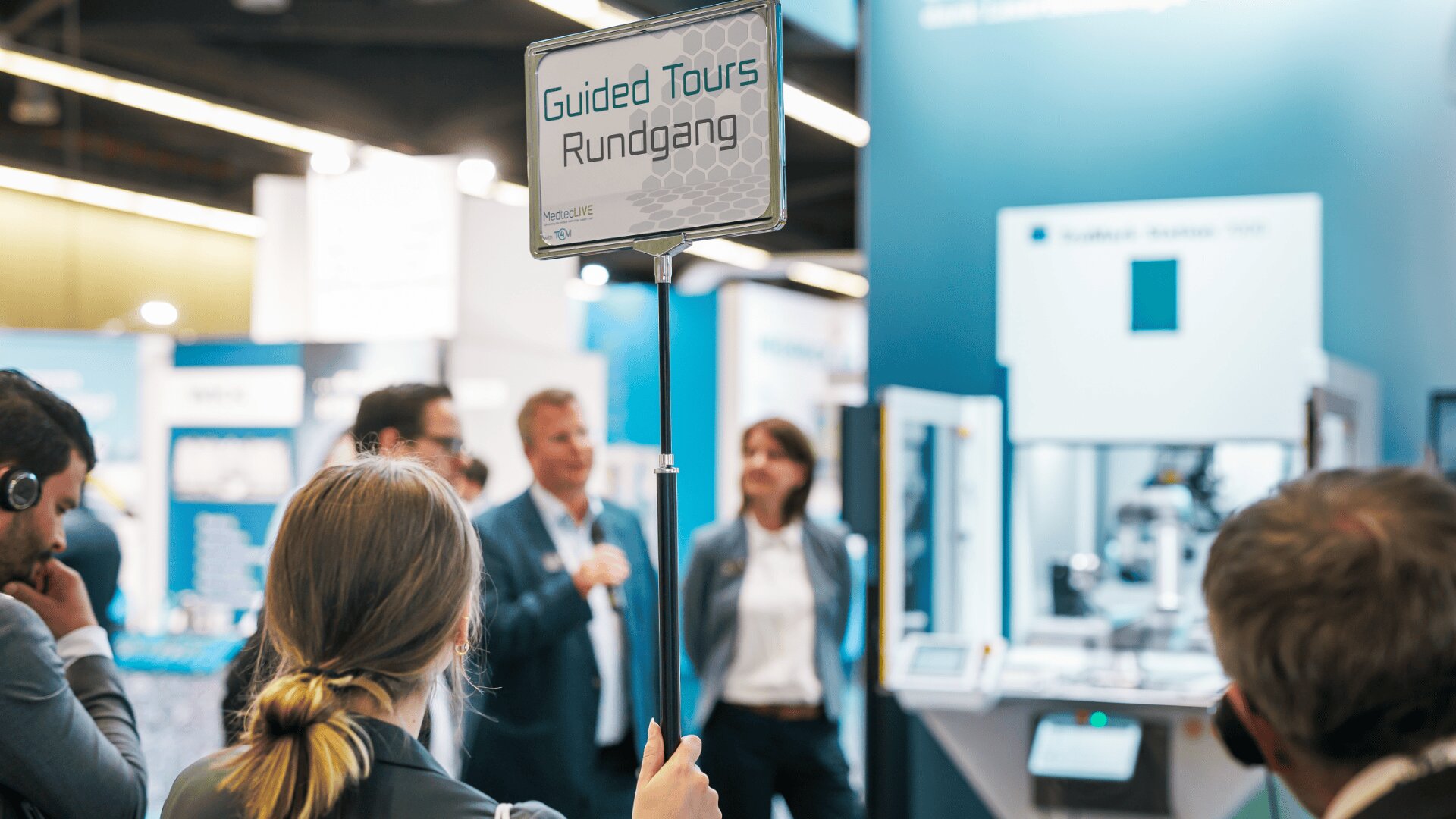 Eine Person hält in einer belebten Ausstellungshalle ein Schild mit der Aufschrift „Guided Tours Rundgang“ hoch. In der Nähe eines Ausstellungsstandes stehen Menschen mit Kopfhörern und hören einem Moderator zu.