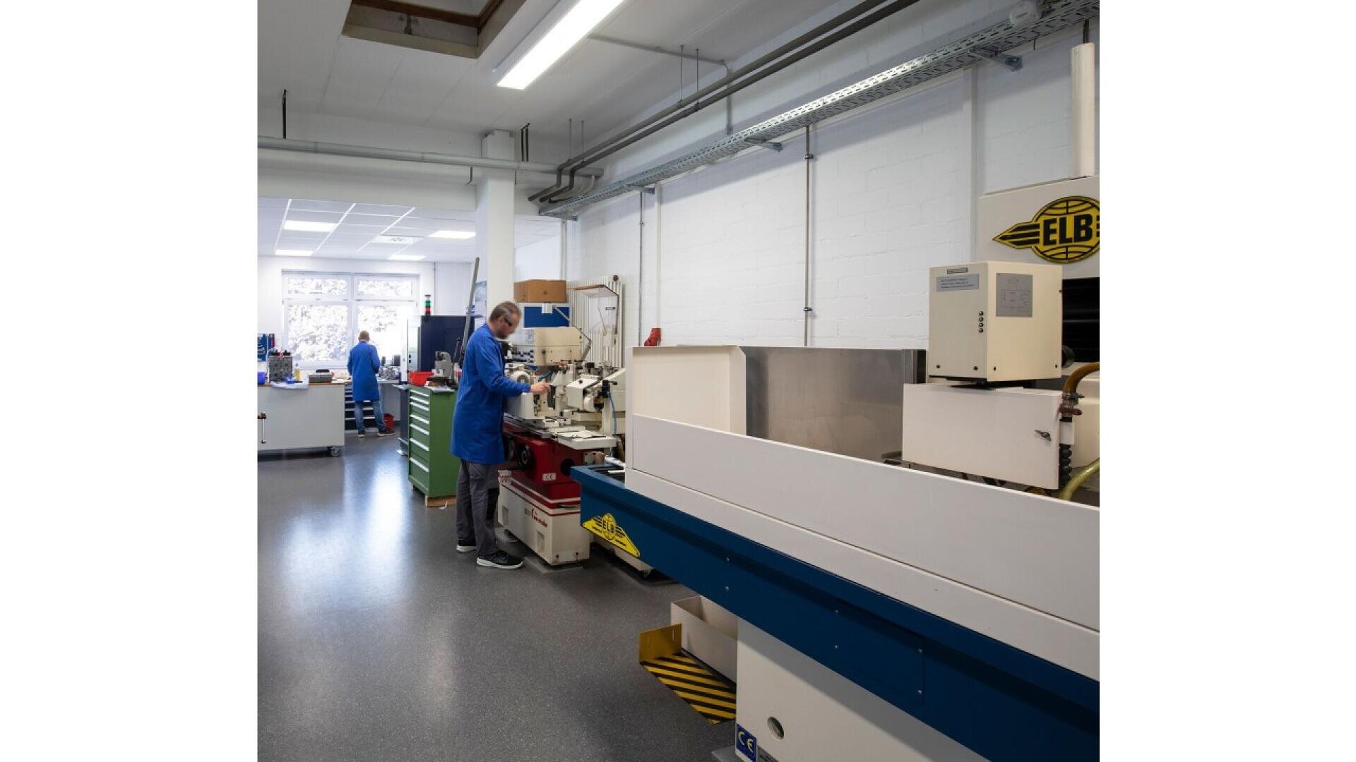 A person in a blue lab coat operates machinery in a clean, well-lit industrial workshop with white walls and various equipment.