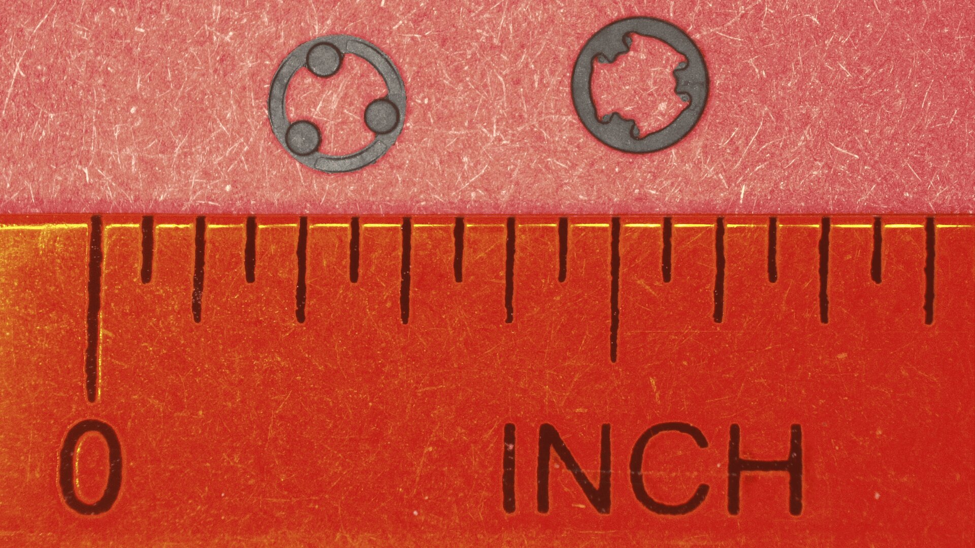 Über einem Lineal mit Zollmaßen sind zwei winzige schwarze Unterlegscheiben mit unterschiedlicher Innenform platziert, wodurch ihre geringe Größe anhand der Maßmarkierungen verdeutlicht wird.