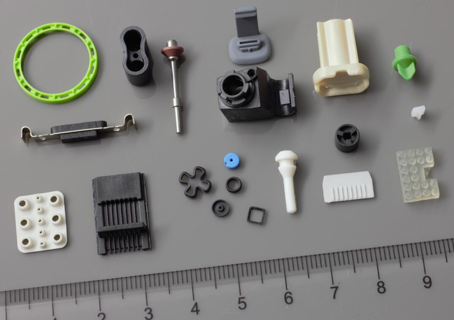 Eine Vielzahl kleiner Kunststoff- und Metallteile sind auf einer grauen Fläche neben einem Lineal angeordnet, das ein Gefühl für den Maßstab vermittelt. Zu den Teilen gehören Zahnräder, Ringe, Verbindungsstücke und andere verschiedene Teile in verschiedenen Farben.