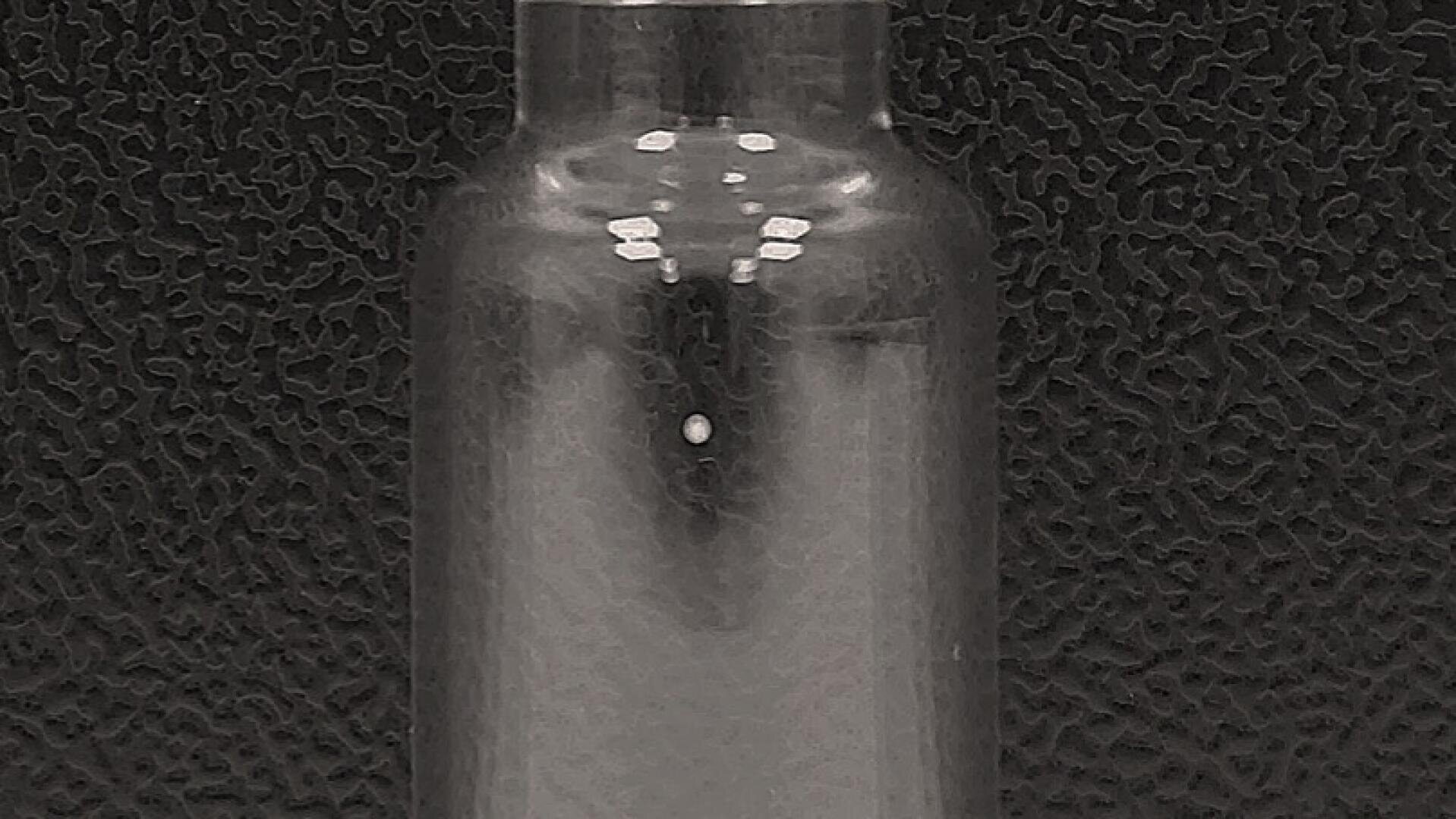 Eine Ampulle aus klarem Glas mit einem silbernen Deckel, die aufrecht vor einem strukturierten schwarzen Hintergrund steht.