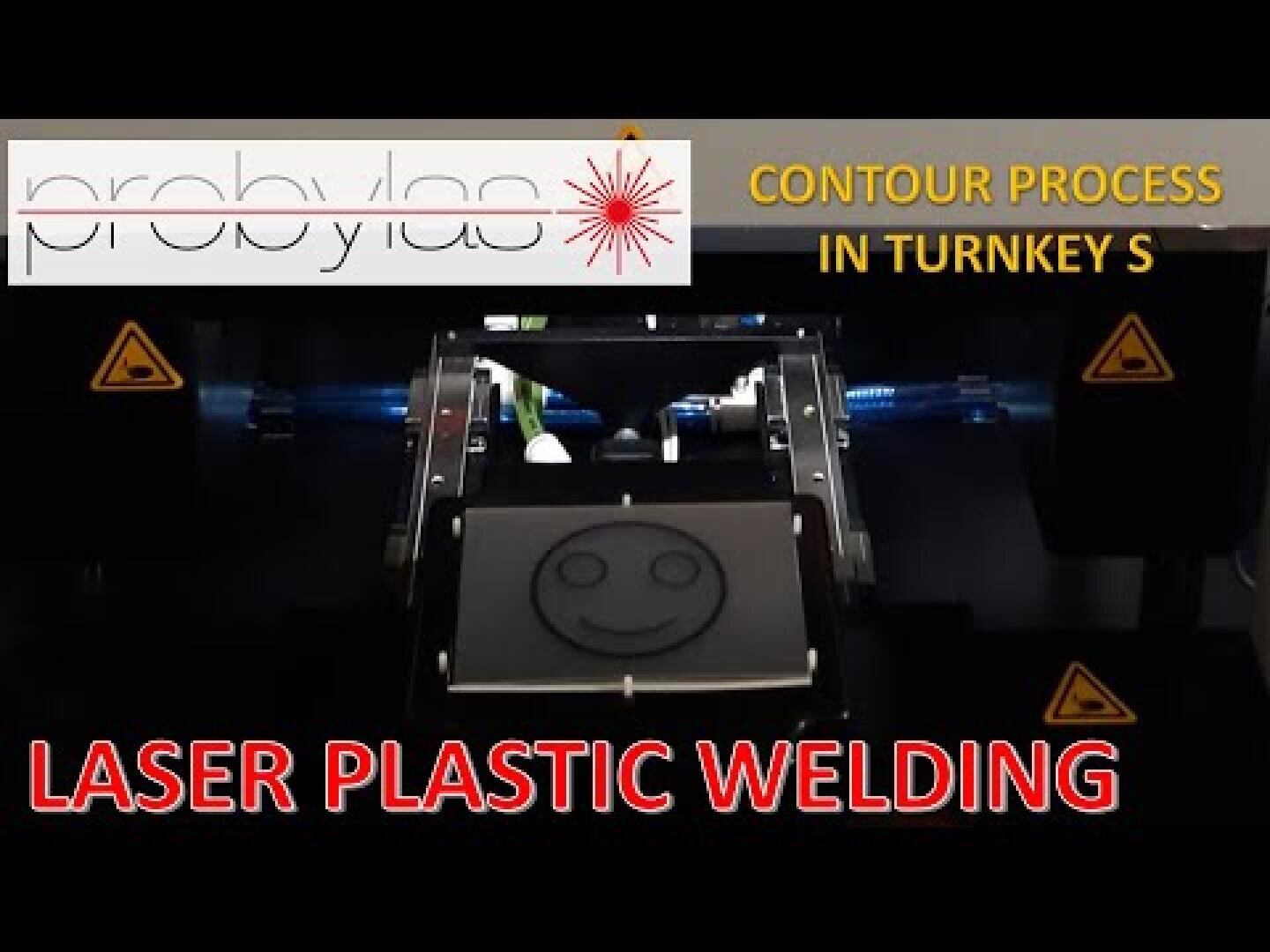A machine that performs laser welding of plastics, with a smiley face on the plastic. The text reads probylas CONTOUR PROCESS IN TURNKEY S and LASER PLASTIC WELDING. Warning signs can be seen on both sides.