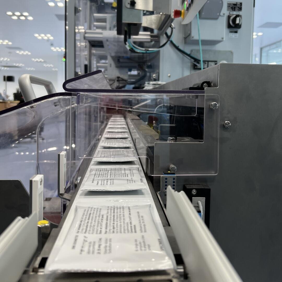 Packages with printed text move along a conveyor belt in a modern, industrial packaging machine, surrounded by metal and clear plastic parts, with bright, evenly distributed ceiling lights in the background.
