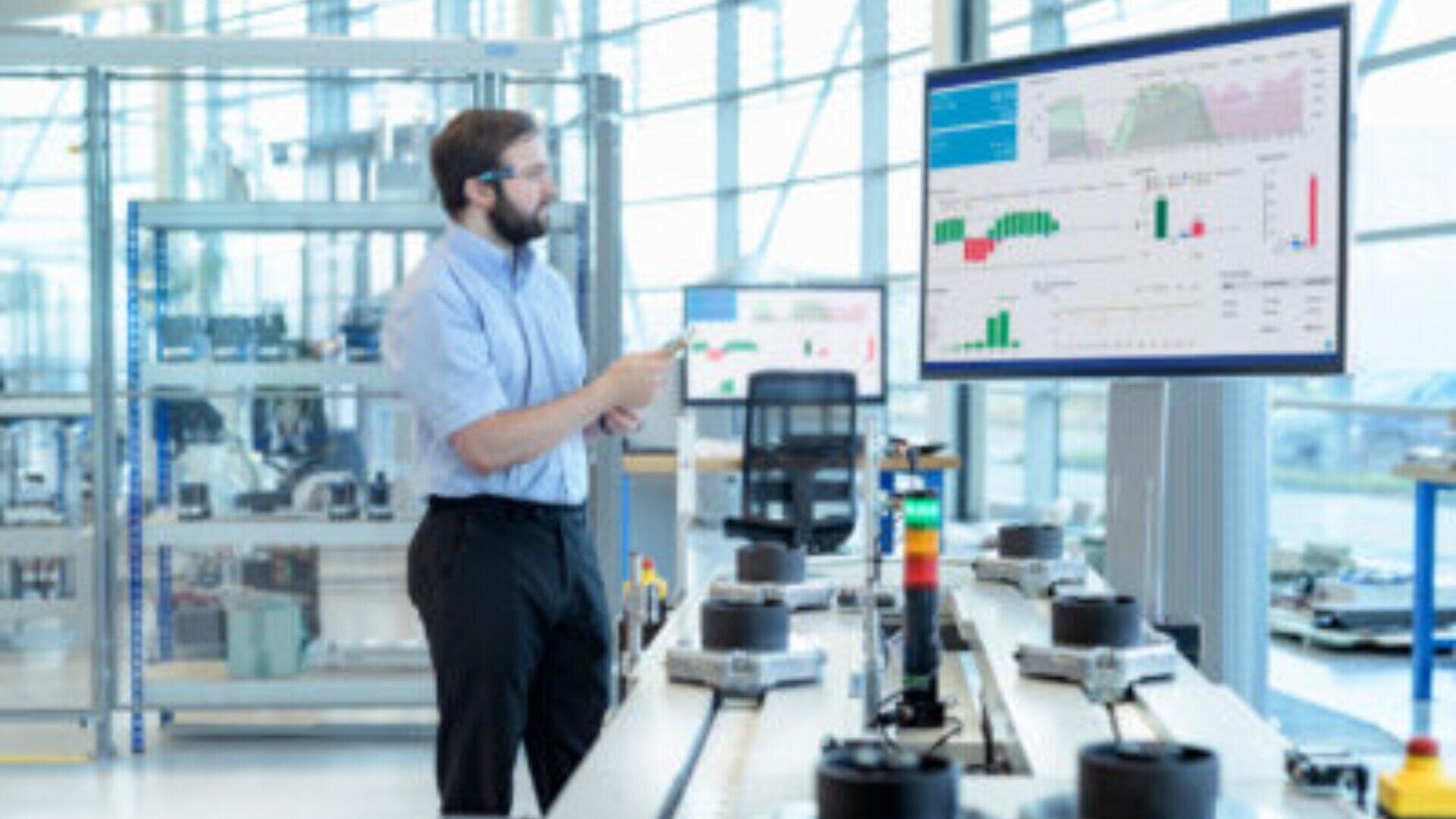 Ein Mann mit Brille und Headset steht in einem modernen Industriearbeitsraum und schaut auf einen großen Monitor, auf dem Diagramme und Daten angezeigt werden. Auf der Werkbank vor ihm sind verschiedene elektronische Geräte und Ausrüstungen angeordnet.