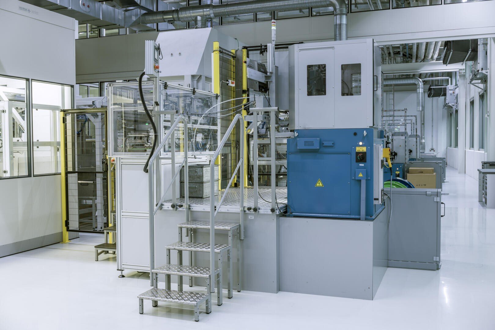 Eine moderne, saubere Industrieanlage mit automatisierten Maschinen, Metalltreppen, Schalttafeln und verschiedenen Geräten, darunter eine große blaue Box, die auf einem polierten weißen Boden steht.