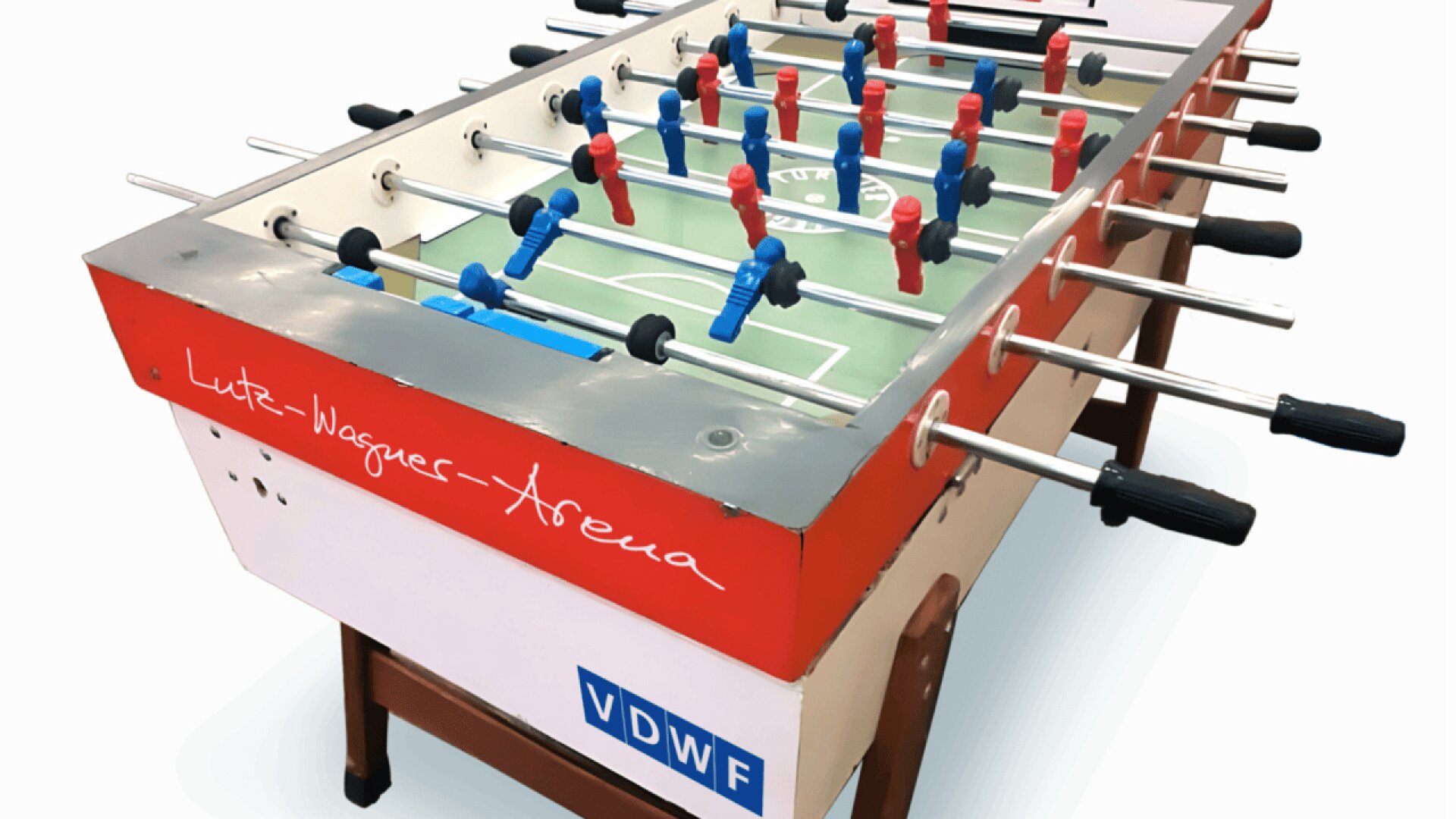 A foosball table with red and blue player figures, black handles, and Lutz-Wagner-Arena written on the side in white script. The table stands on brown legs and has a VDW logo on the front.