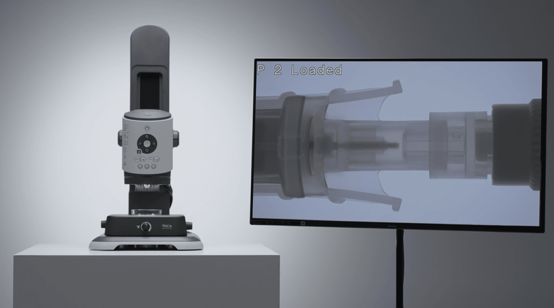 A robotic medical device stands on a platform next to a monitor that shows a close-up of the mechanism of a syringe or vial and illustrates precise loading or alignment.