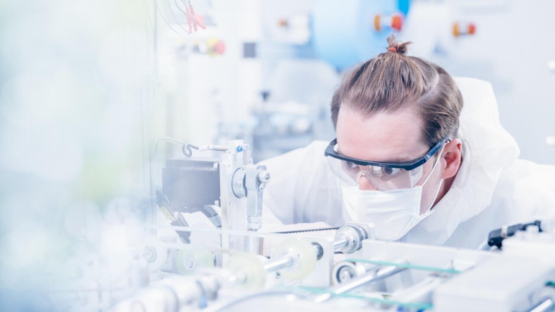 A scientist wearing safety goggles, a face mask, and protective clothing examines equipment in a bright, modern laboratory.