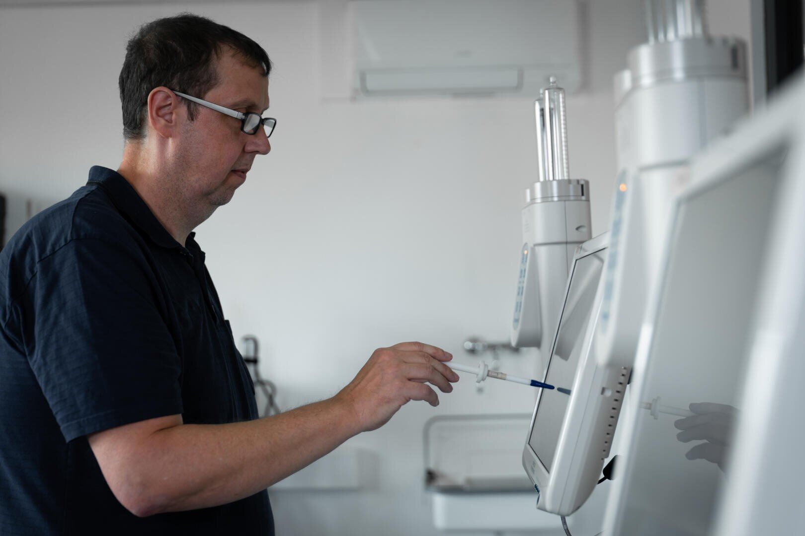 A man wearing glasses interacts with a touchscreen medical device in a clinical setting by using a stylus while standing next to other medical equipment.
