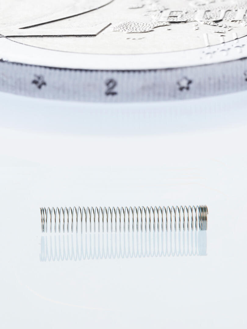 A small, tightly coiled metal spring is placed on a reflective surface with part of a coin in the background to show the size comparison and intricate details of the spring coils.