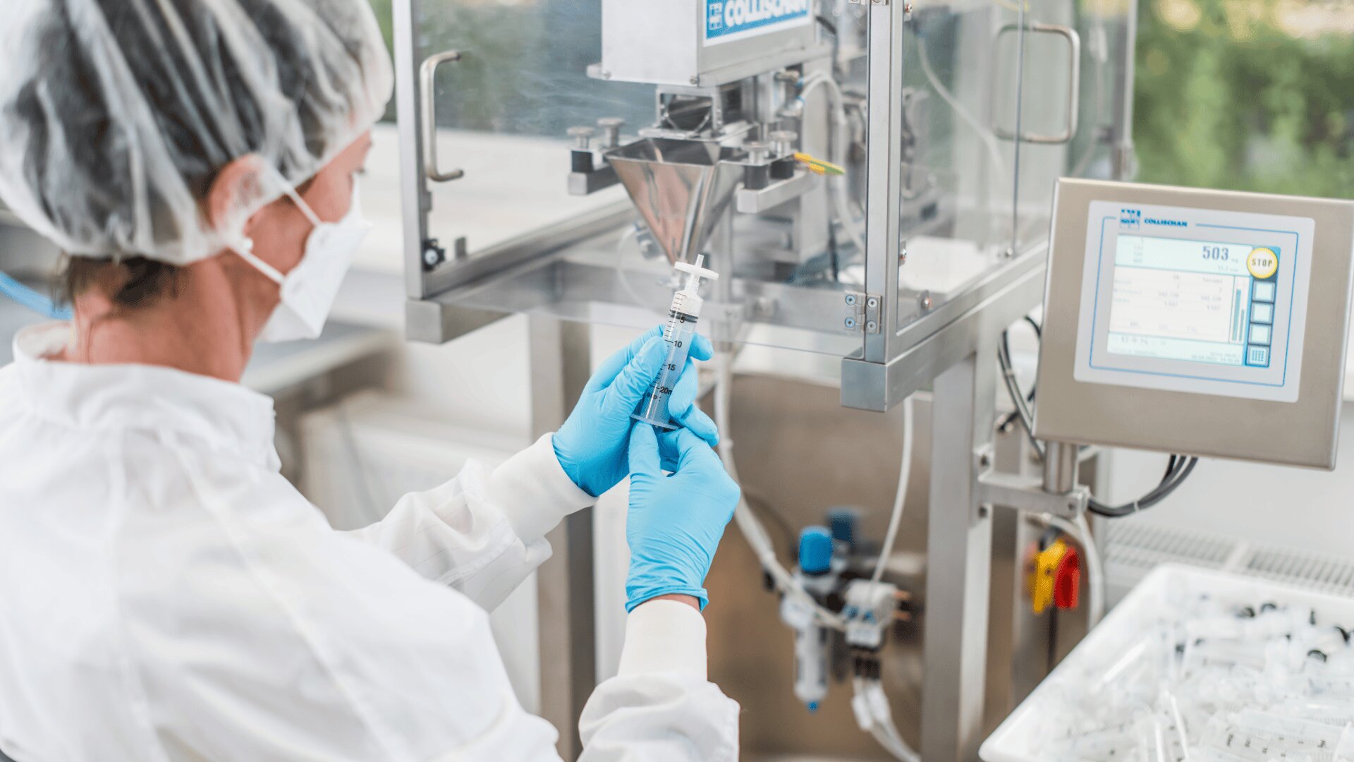 A person in protective clothing, mask, and gloves fills a syringe with liquid from a machine in a laboratory or cleanroom, with trays of syringes and a digital display visible nearby.
