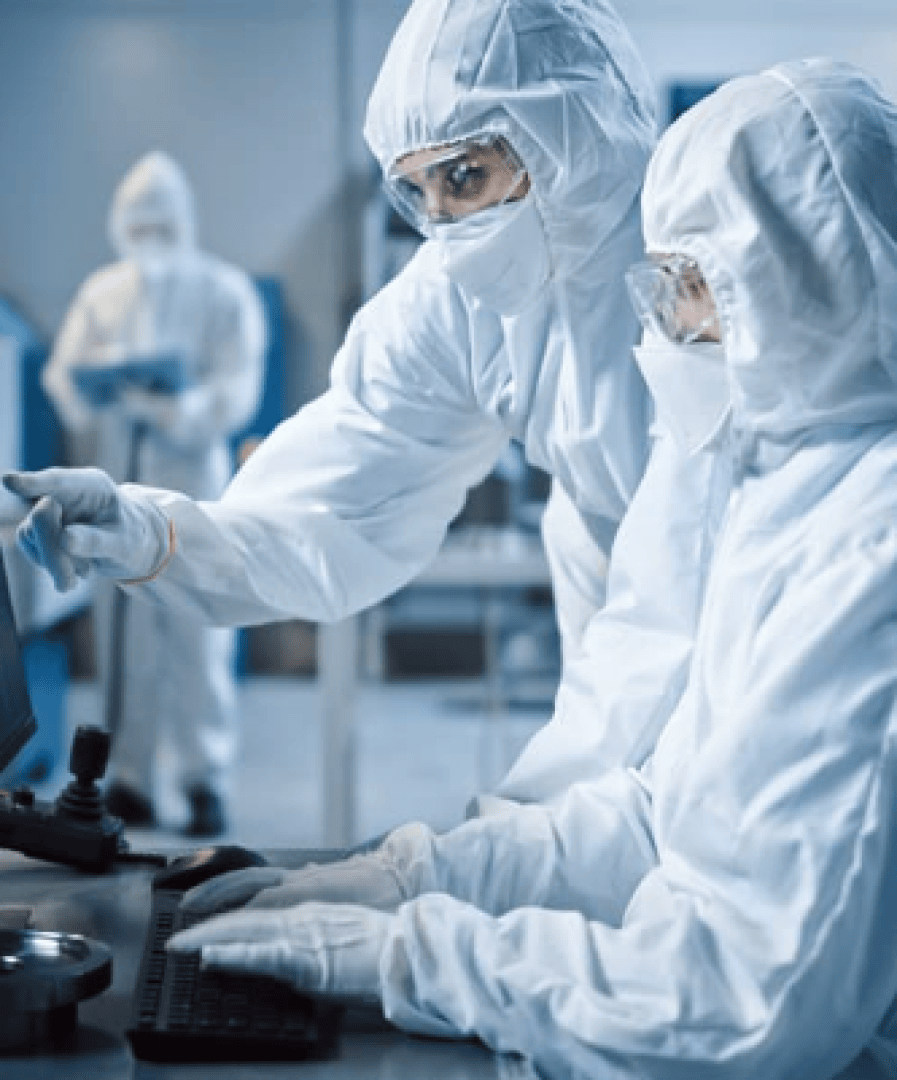 Two scientists in full protective suits and goggles work together on a computer in a laboratory, one pointing at the screen and another person can be seen out of focus in the background.
