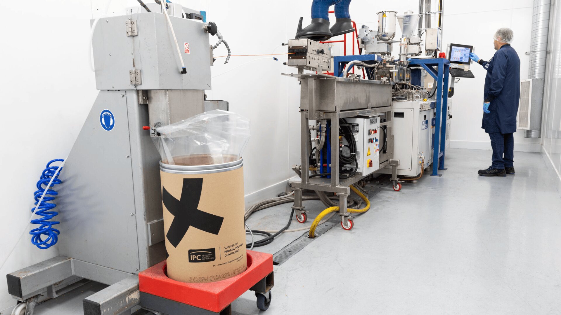 A person in a blue coat operates a machine in a clean, industrial setting with various equipment and cables. In the foreground, a large cardboard drum lined with plastic sits on a red cart.