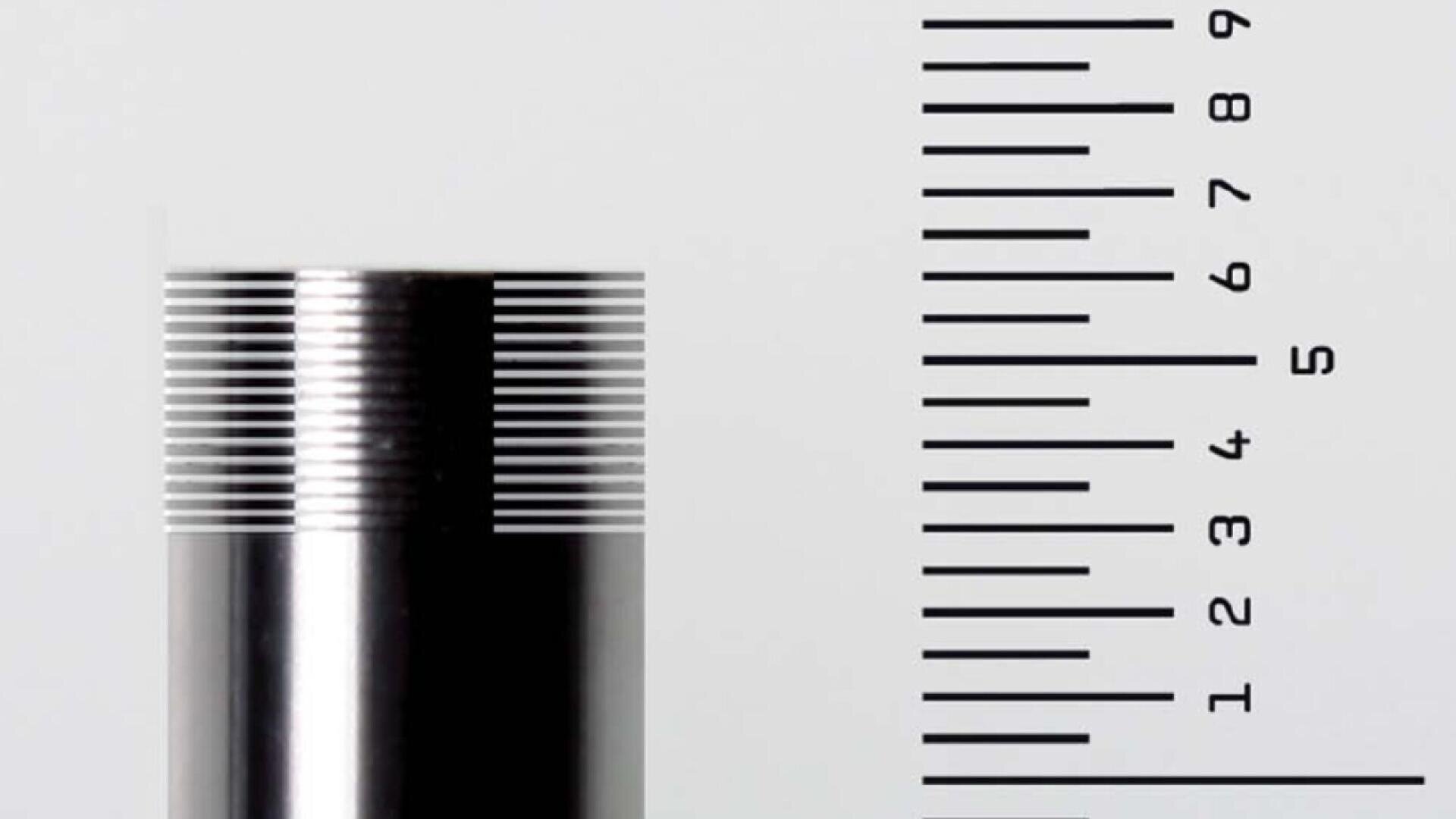 A close-up of a metal rod with fine, evenly spaced grooves next to a vertical ruler marked from 0 to 9, illustrating precision measurement.