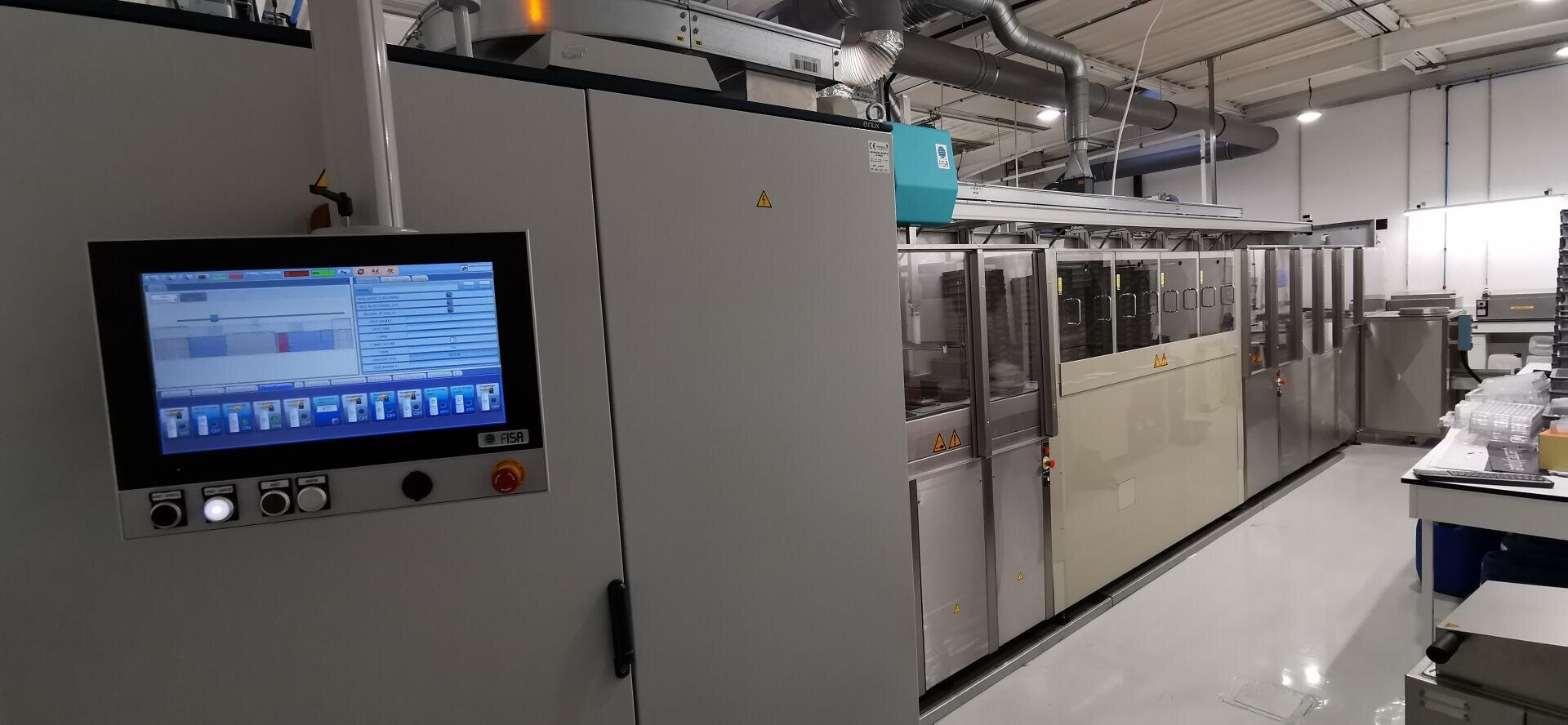 Ein modernes industrielles Bedienfeld mit Touchscreen-Oberfläche ist auf einer großen grauen Maschine in einer sauberen, hell erleuchteten Fabrikumgebung montiert. Im Hintergrund sind weitere automatisierte Geräte zu sehen.