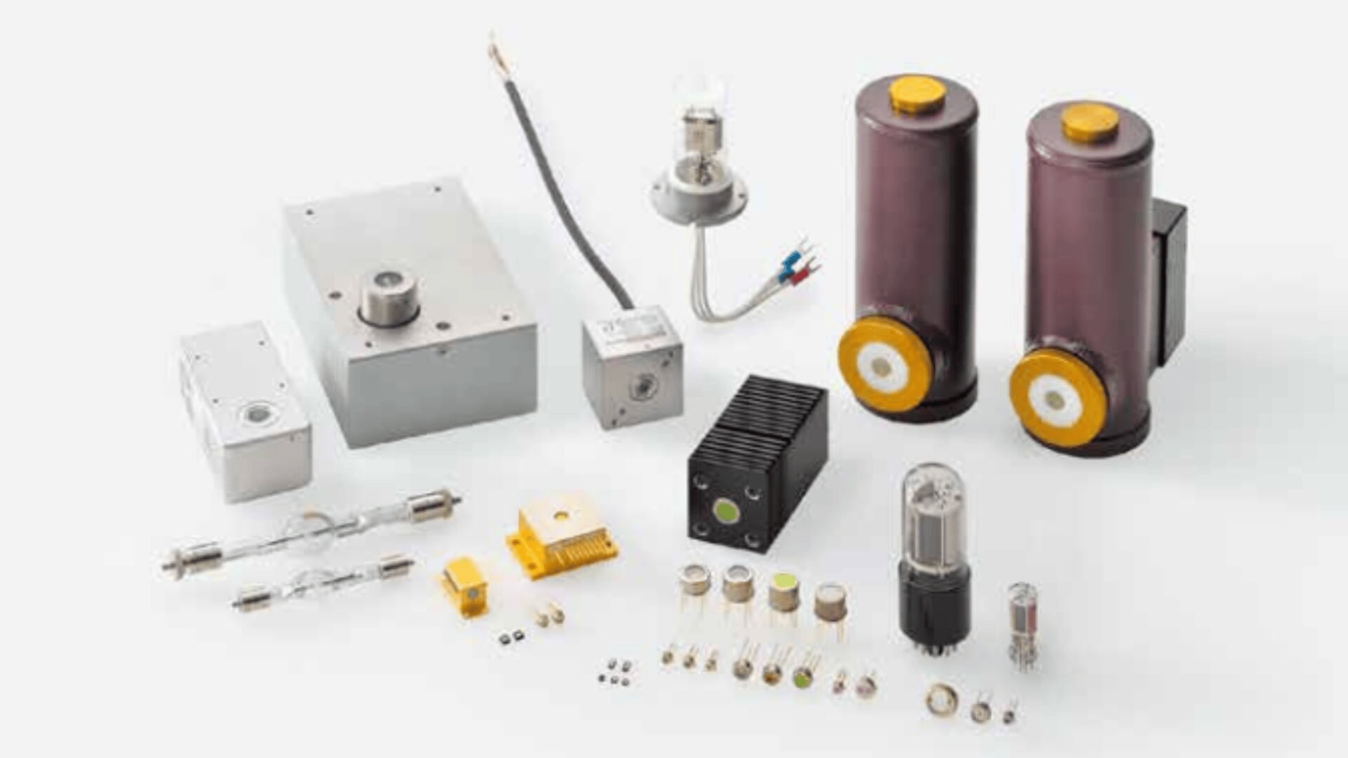 Eine Auswahl elektronischer Komponenten und Geräte, darunter Vakuumröhren, zylindrische Kondensatoren, Leiterplatten, Steckverbinder und verschiedene Kleinteile, ordentlich auf einer weißen Oberfläche angeordnet.