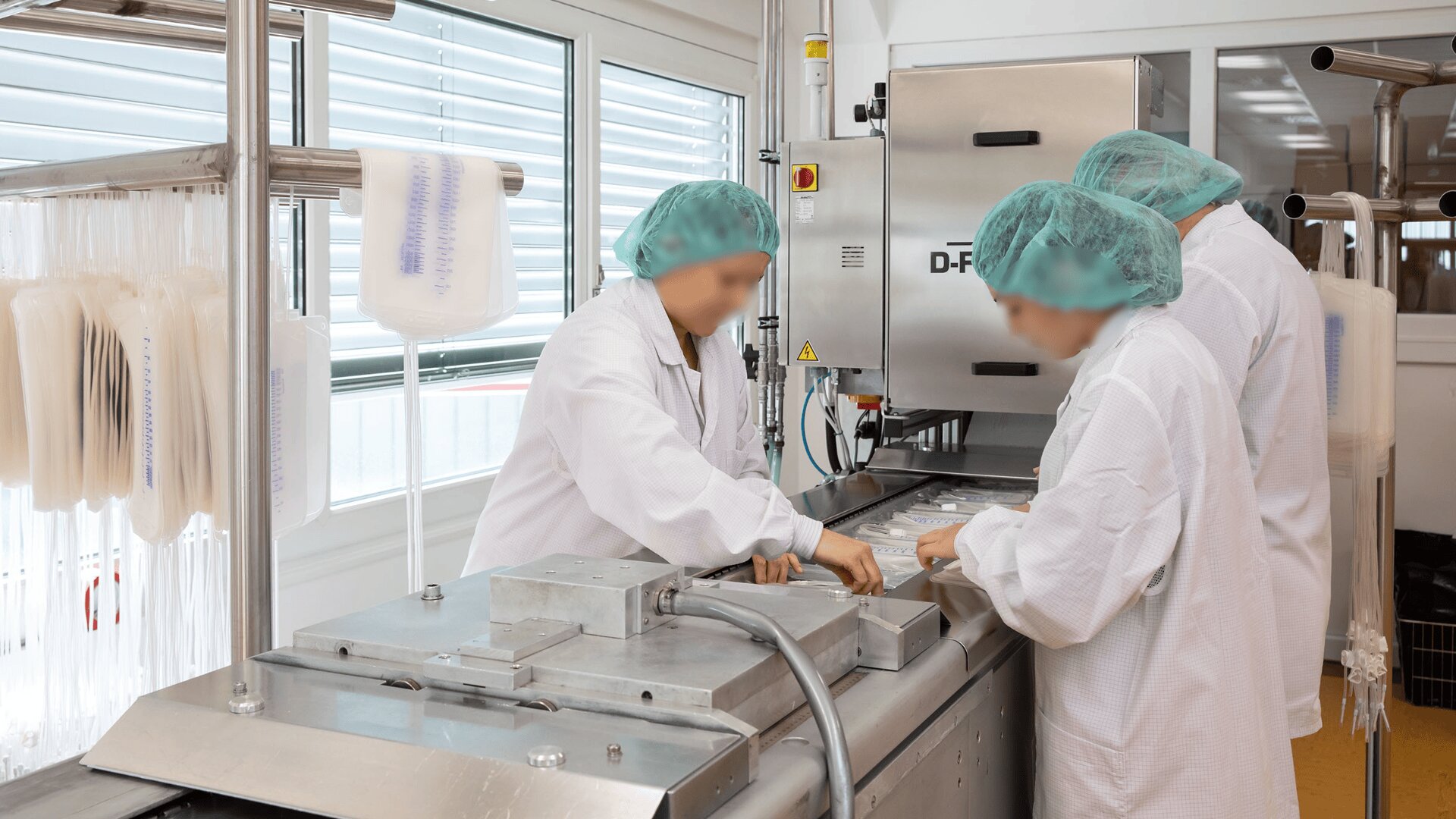 Drei Personen in weißen Laborkitteln, Haarnetzen und Masken arbeiten gemeinsam an einer Edelstahlmaschine in einem sauberen, hell erleuchteten Labor oder einer Lebensmittelverarbeitungsanlage. Plastiktüten hängen an einem Gestell in der Nähe.