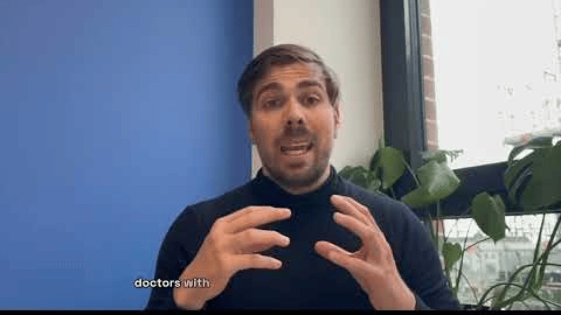 A man with short dark hair and a beard, wearing a black turtleneck, speaks animatedly with hands raised in front of a blue wall and window with green plants. Subtitles partially read, doctors with.