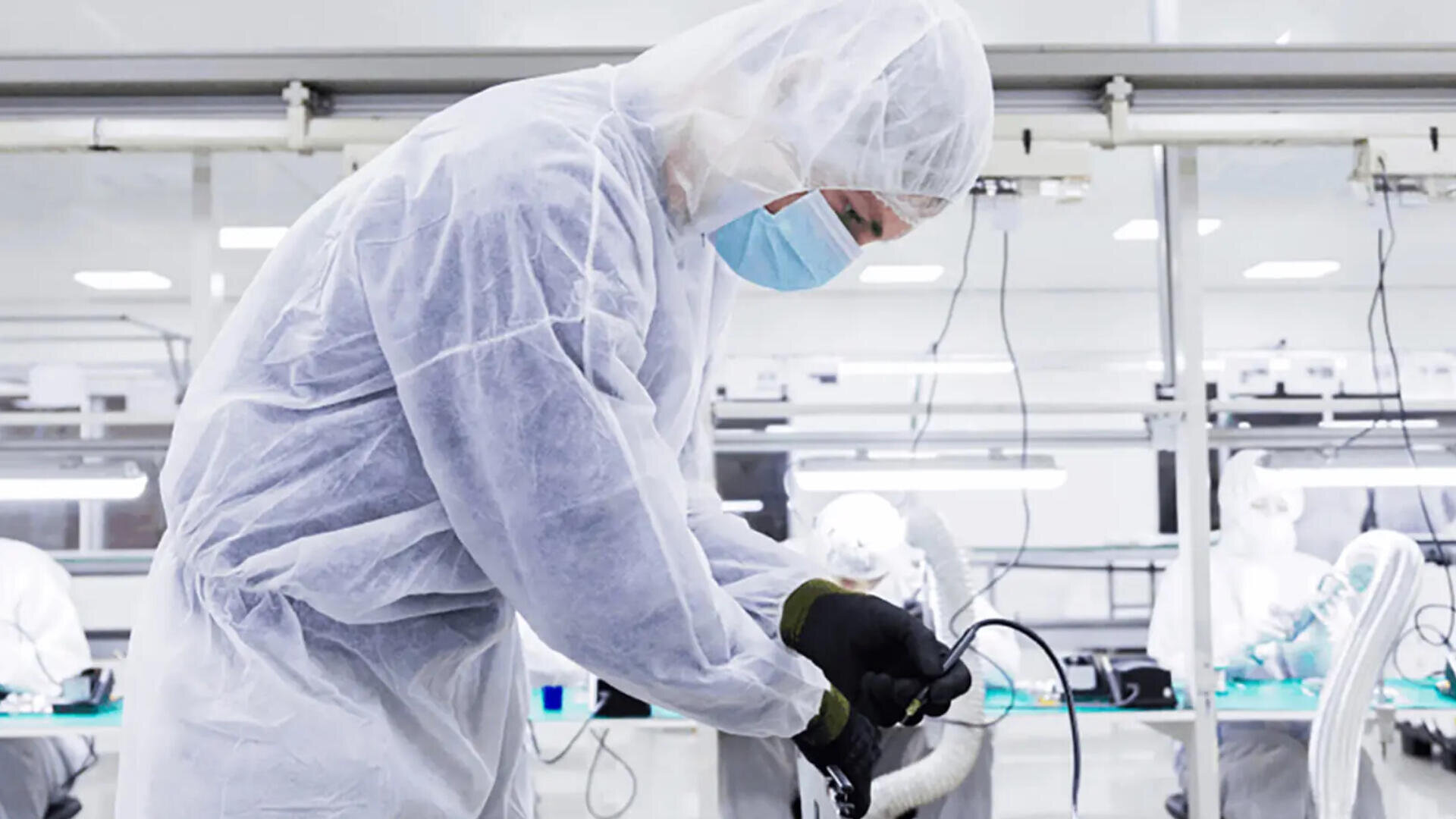 Eine Person in Schutzkleidung (Maske und Handschuhe) arbeitet mit elektronischen Geräten in einem sauberen Hightech-Labor oder einer Produktionsumgebung. Im Hintergrund sind weitere Arbeiter zu sehen.