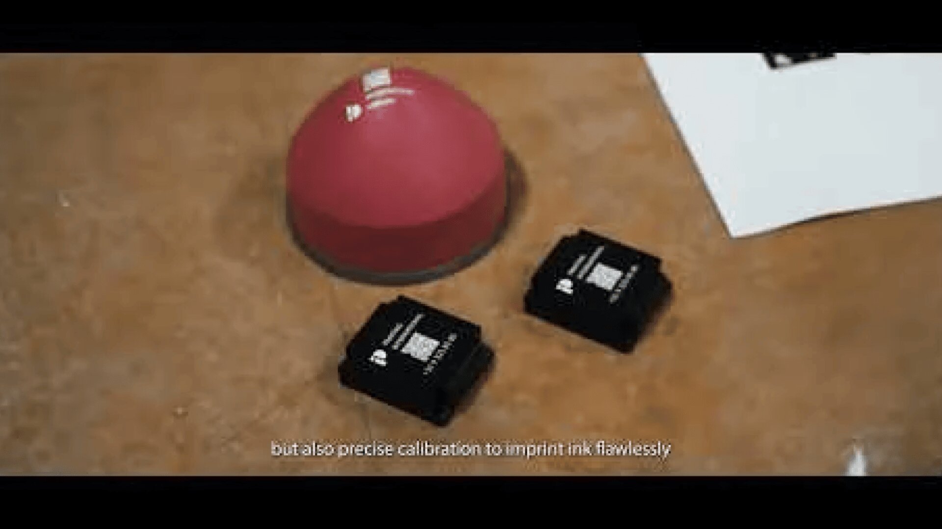 A red dome-shaped object and two small black electronic devices are placed on a brown surface, with a piece of white paper partially visible in the corner. Subtitles at the bottom read: “but also precise calibration to imprint ink flawlessly”.