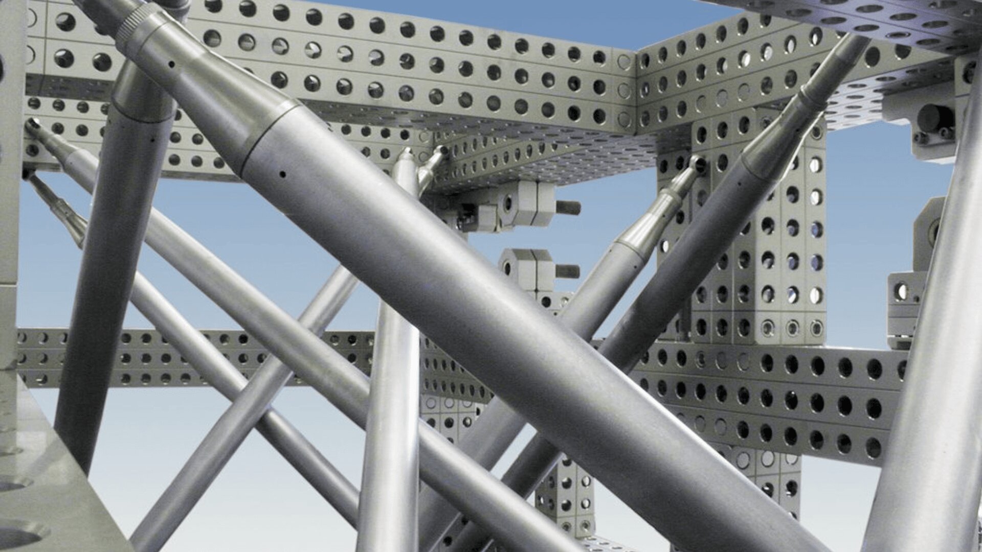 Eine Nahaufnahme eines metallischen Rahmens mit perforierten Balken und sich kreuzenden zylindrischen Stäben, die vor dem Hintergrund eines blauen Himmels eine geometrische Struktur bilden.