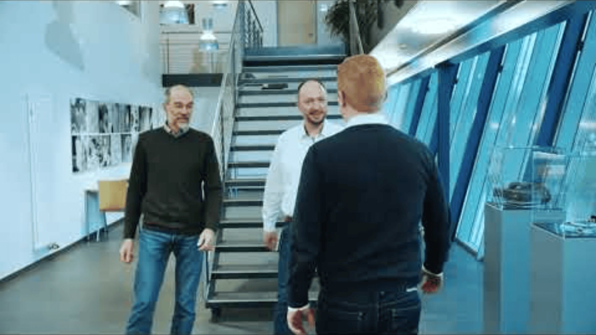 Three men are standing and talking near a modern staircase inside a bright office building with large windows and photographs on the wall.