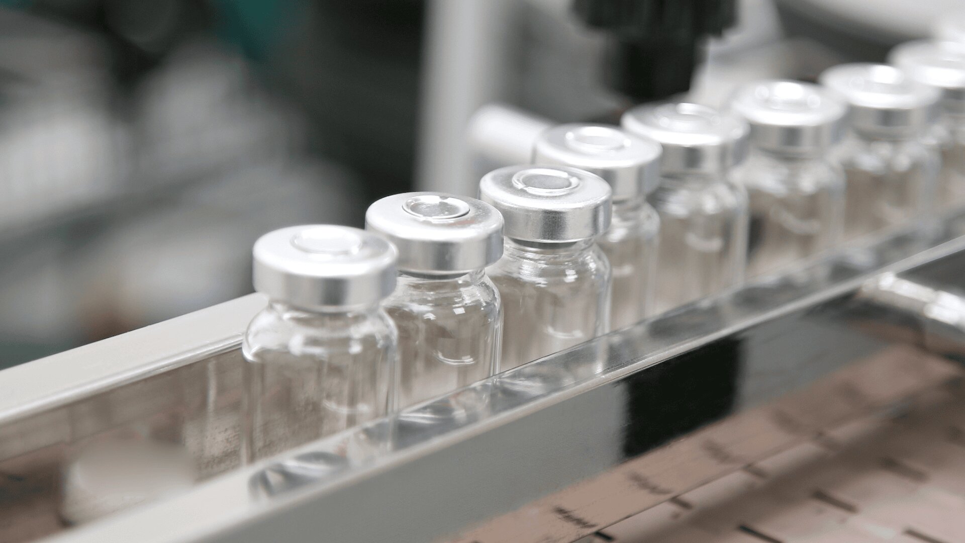 Eine Reihe durchsichtiger Glasfläschchen mit silbernen Verschlüssen ist auf einem Metallproduktionsband aufgereiht und lässt auf eine pharmazeutische oder Laborumgebung schließen.