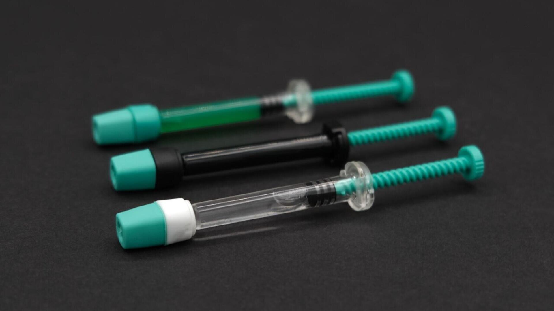 Three syringes with green plungers and teal caps are arranged on a dark surface. One syringe contains a green liquid, one is empty and transparent, and one is black.