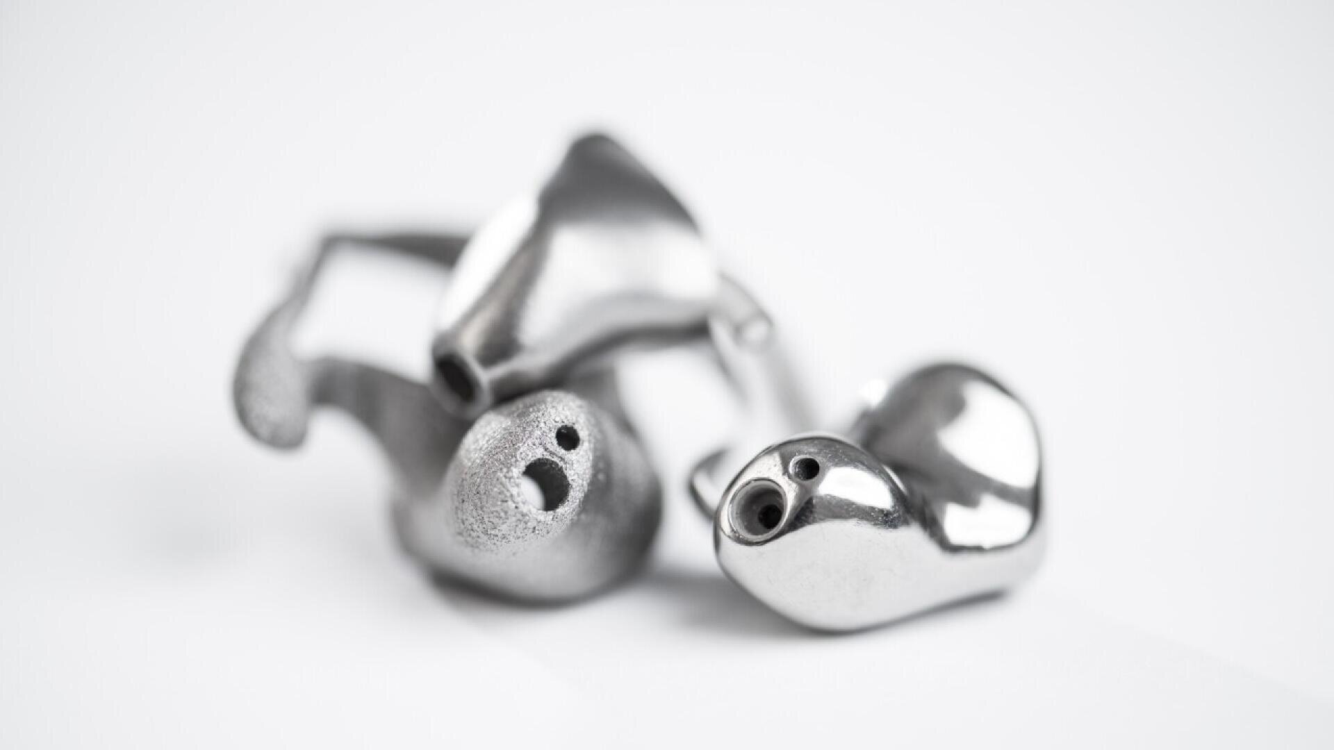 Two metallic custom-molded in-ear monitors are placed on a white surface. One earpiece has a matte finish, while the other appears shiny and polished. Both have visible sound bores and a smooth, curved design.