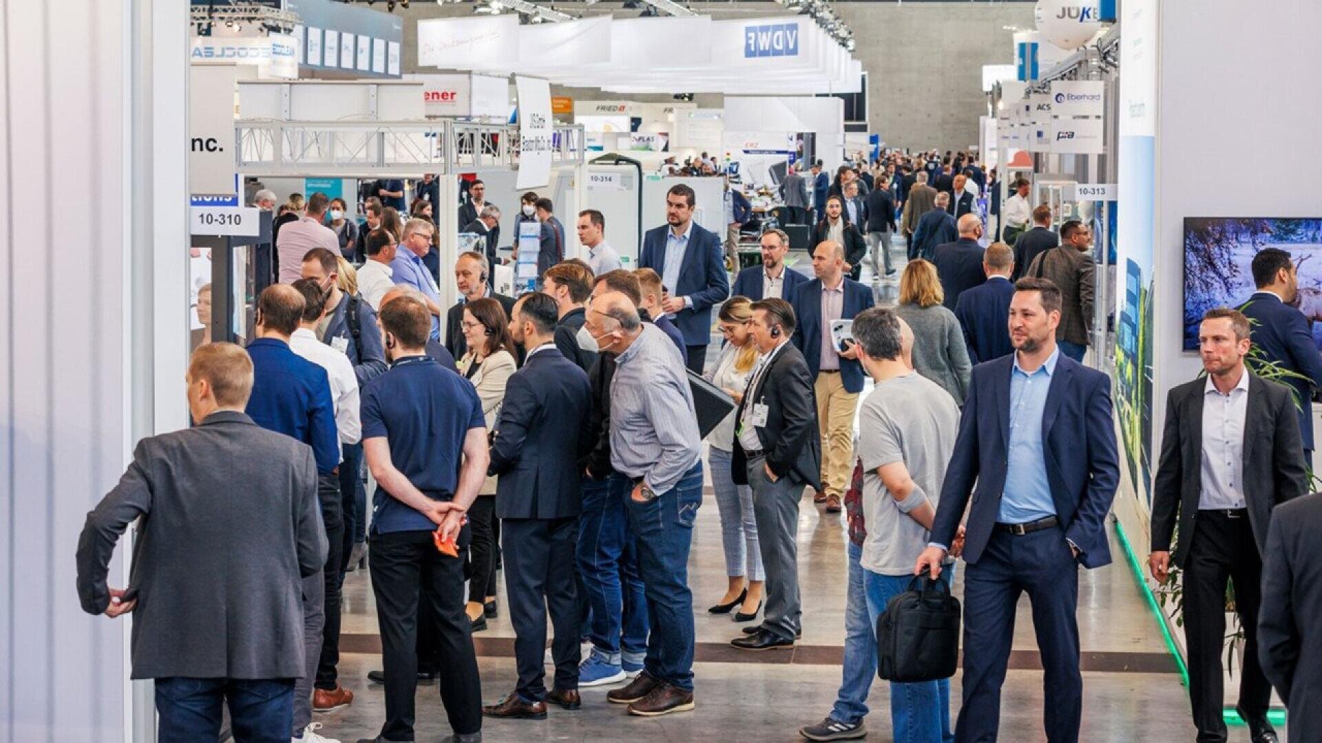 A busy convention hall filled with people in business attire walking, talking, and interacting among various booths and displays at a trade show or expo.