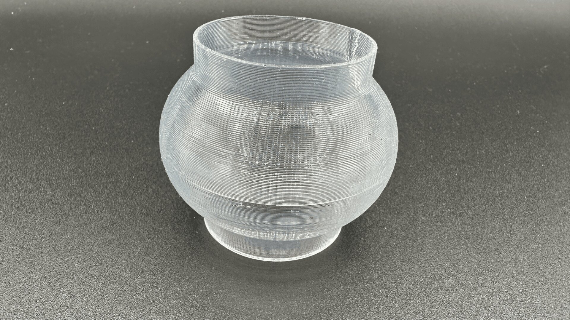 Eine klare, runde, gerippte Kunststoffvase mit breitem Körper und schmaler Öffnung, platziert auf einer dunklen, strukturierten Oberfläche.