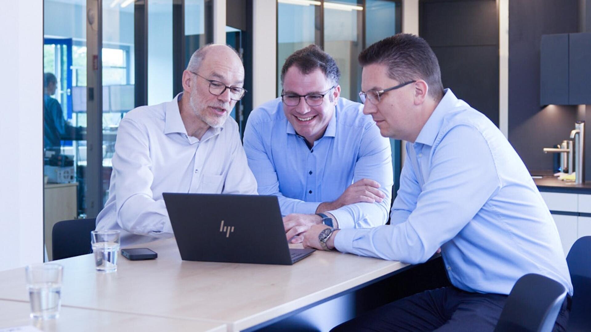 Drei Männer im Geschäftsanzug sitzen an einem Tisch, schauen auf einen Laptop und lächeln. Zwei Wassergläser stehen auf dem Tisch, und im Hintergrund ist ein modernes Büro mit großen Fenstern zu sehen.