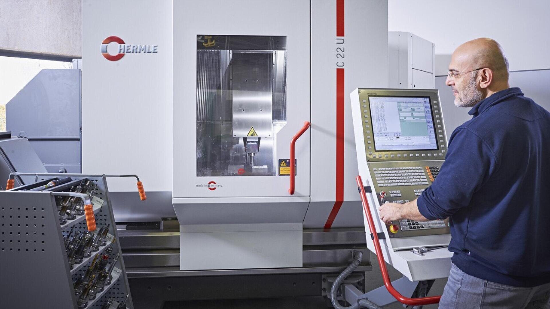 A man operates a CNC machine using a control panel in a modern industrial setting, with tools and equipment visible nearby. The CNC machine is branded Hermle C 22 U.