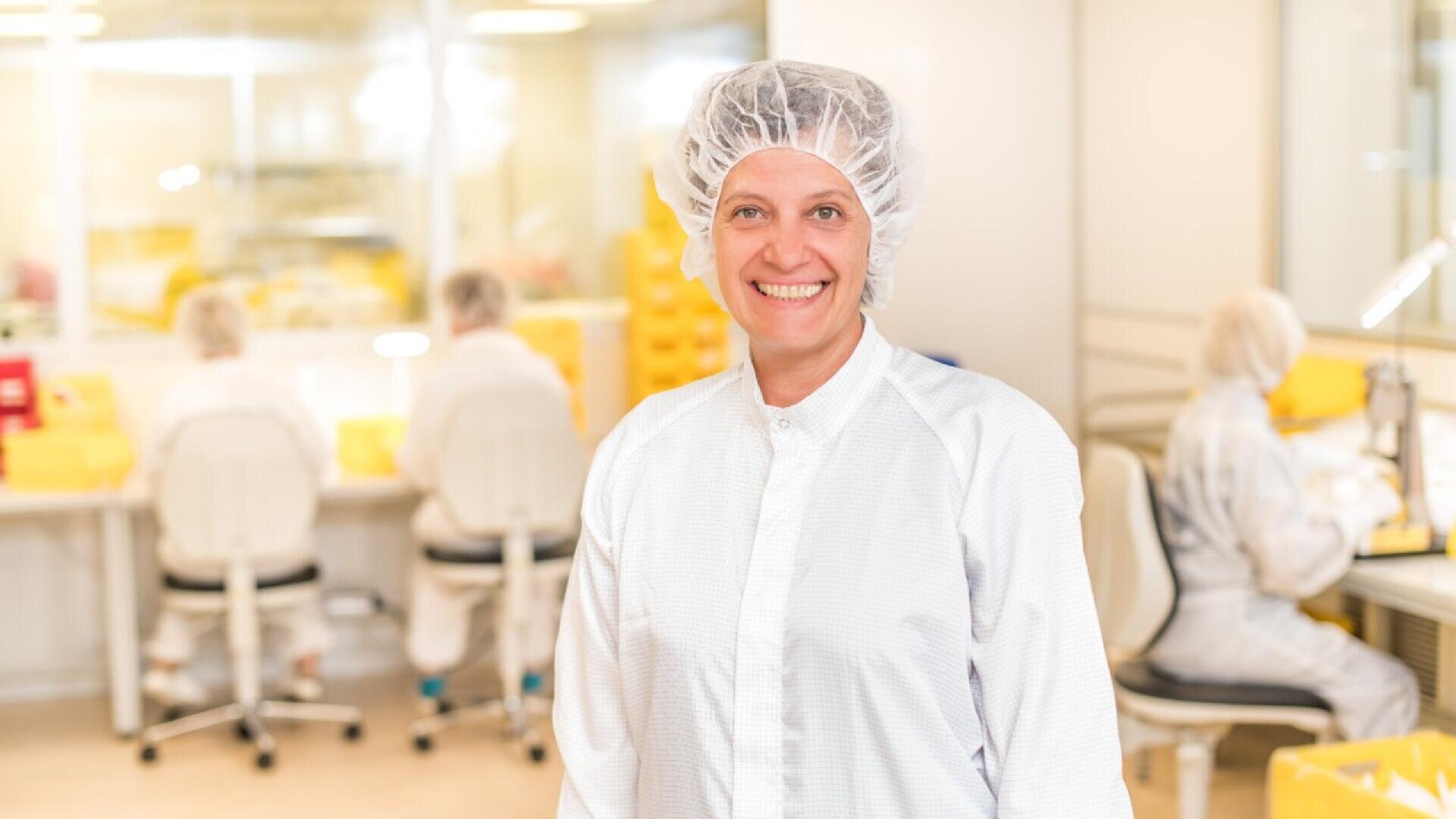 A smiling woman in a white lab coat and hairnet stands in a bright laboratory, while several people in similar attire work at desks in the background.