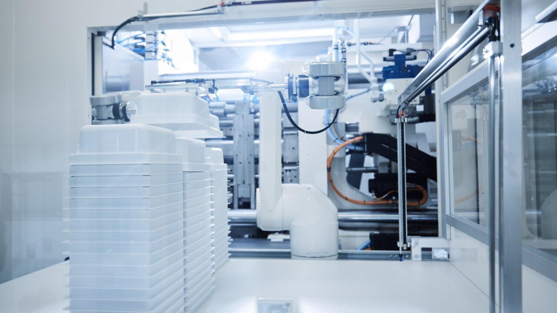 A modern automated factory scene with white plastic containers stacked in the foreground and robotic machinery operating in the background under bright lighting.