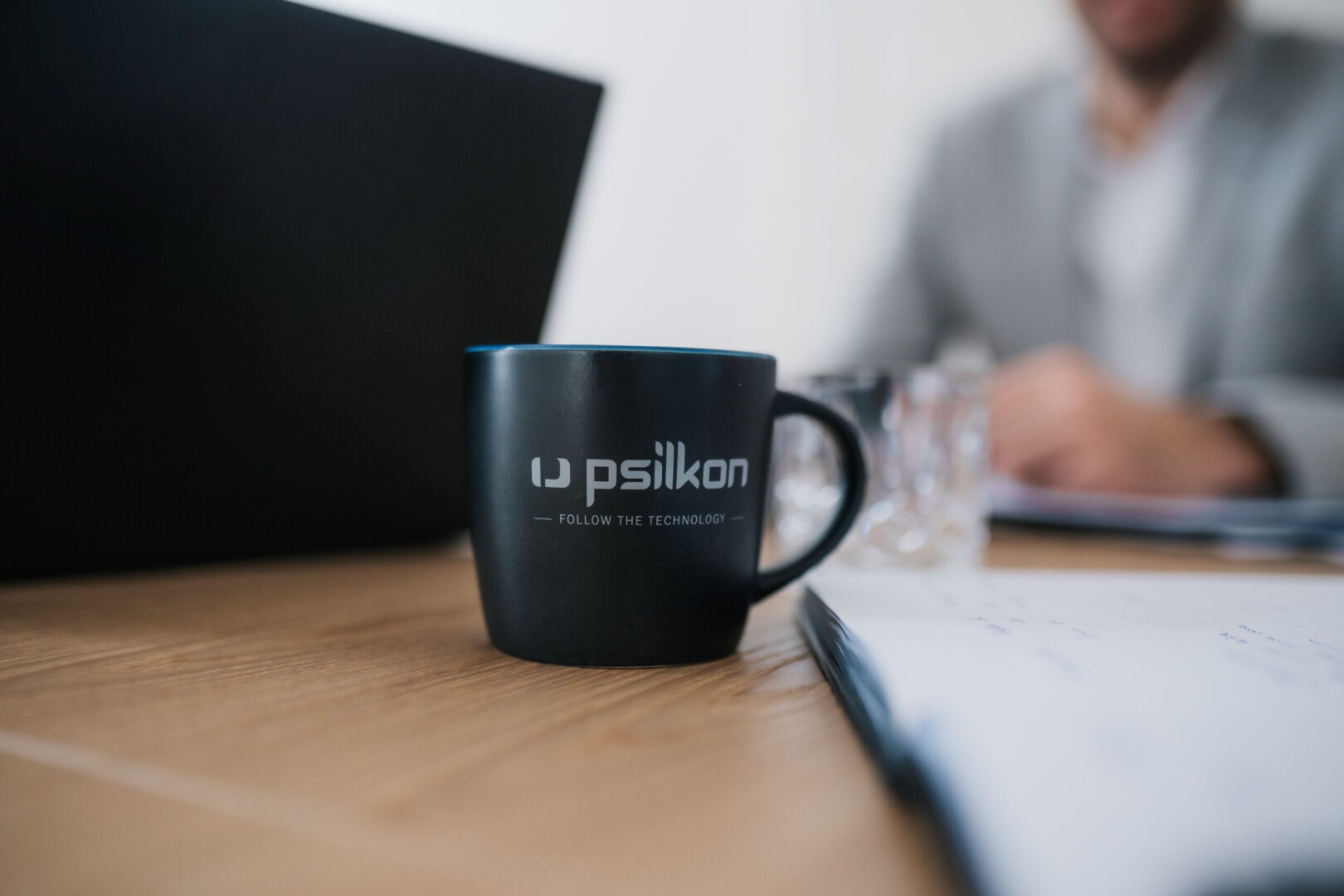 Eine schwarze Tasse mit dem Logo und dem Text „Psilkon FOLLOW THE TECHNOLOGY“ steht auf einem Holzschreibtisch neben einem Laptop, Papieren und einer verschwommenen Person im Anzug im Hintergrund.