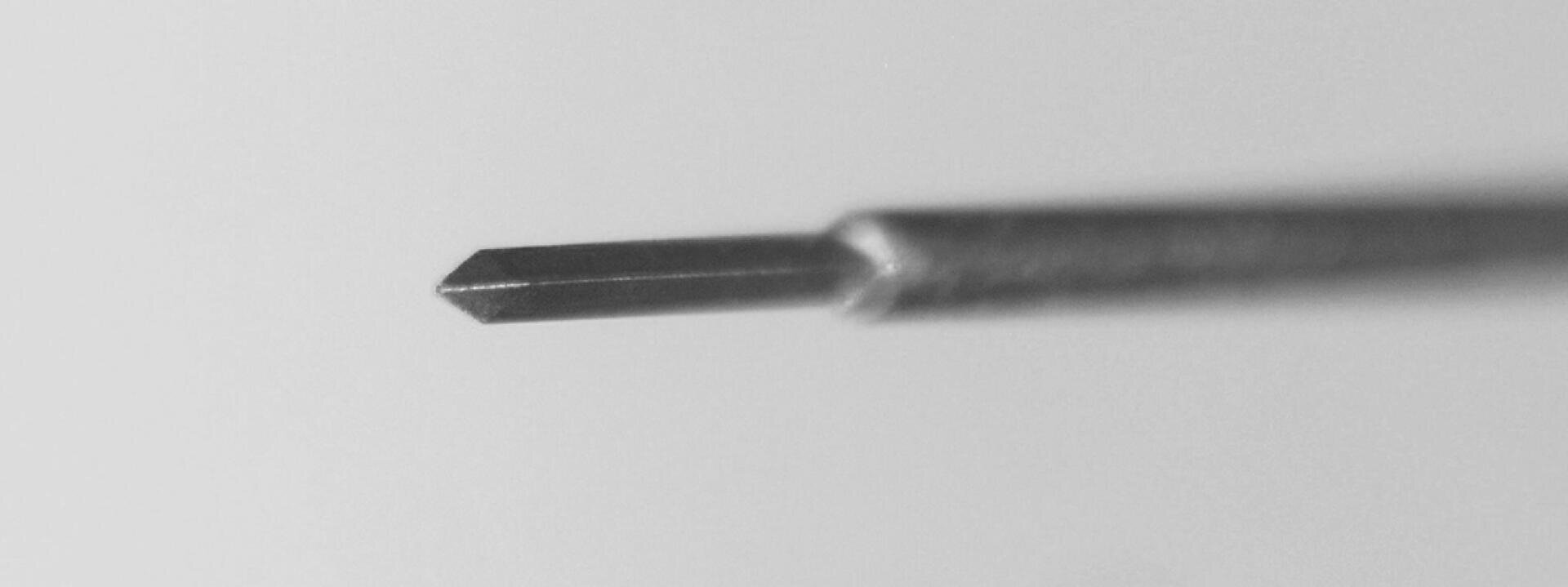 Close-up of a small metal tool with a pointed, chiseled tip, reminiscent of a precision screwdriver or similar fine-tipped instrument, against a plain, blurred background.
