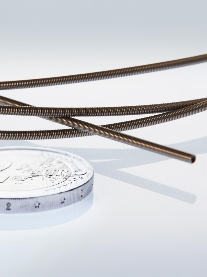 Close-up of a silver coin next to several thin, coiled metal wires arranged diagonally on a reflective white surface. The coin provides a sense of scale for the wires.