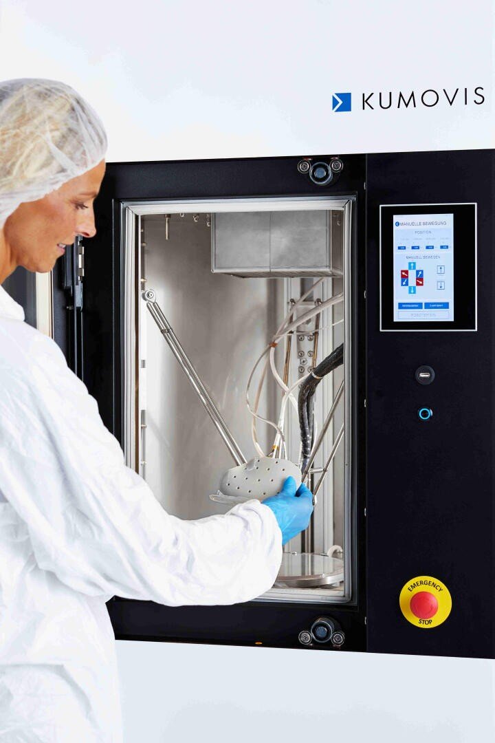 Eine Person in Schutzkleidung und Handschuhen bedient einen Kumovis 3D-Drucker mit Touchscreen und hält ein teilweise gedrucktes Objekt in der Maschine.