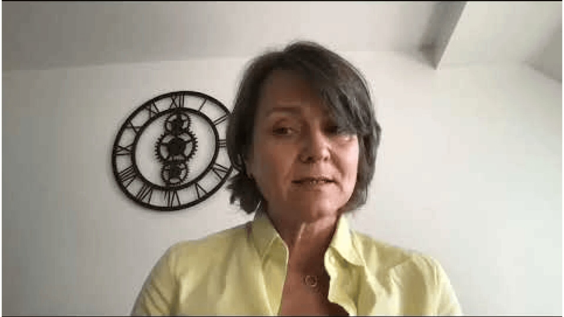Eine Frau mit kurzen braunen Haaren und einem gelben Hemd sitzt drinnen vor einer weißen Wand, an der eine große schwarze Uhr hängt. Sie scheint während eines Videoanrufs zu sprechen oder zuzuhören.