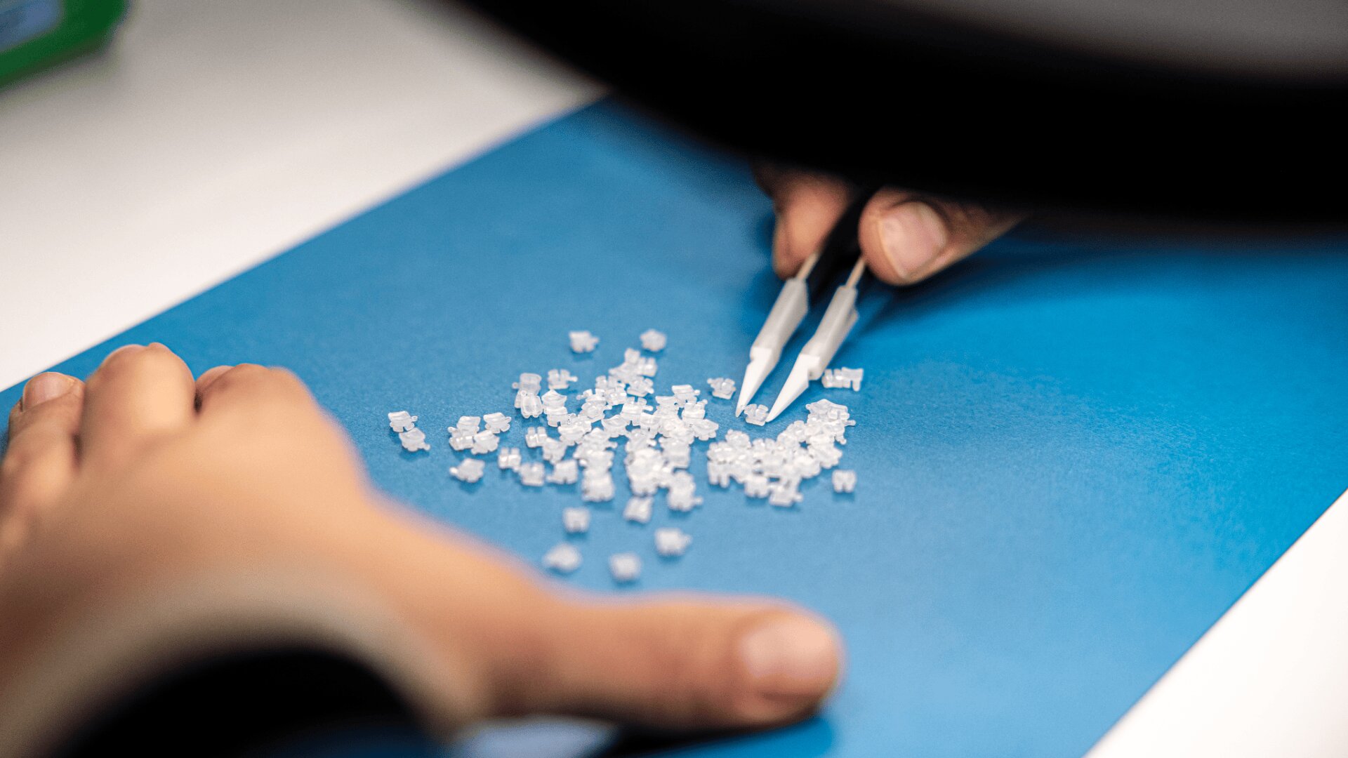 A person uses white tweezers to arrange or inspect small white plastic pellets spread on a blue surface, with another hand steadying the paper.