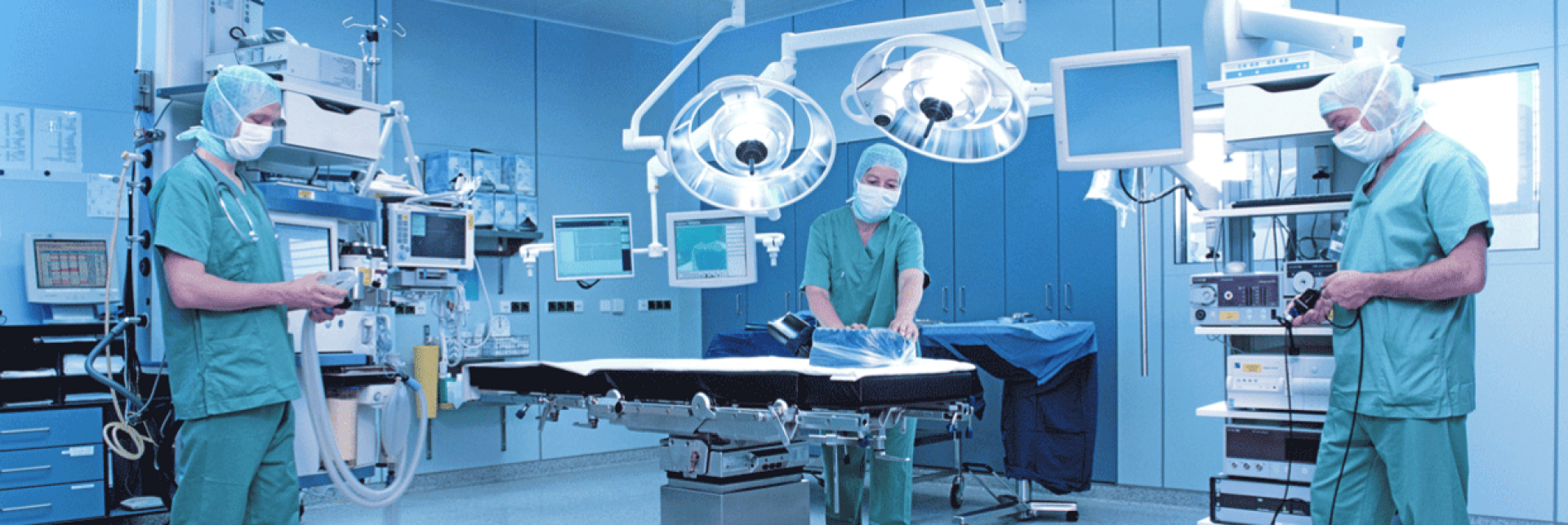 Three medical professionals in gowns and masks stand in a modern, well-lit operating room equipped with surgical lights, monitors, and medical equipment, preparing for a procedure around an empty operating table.