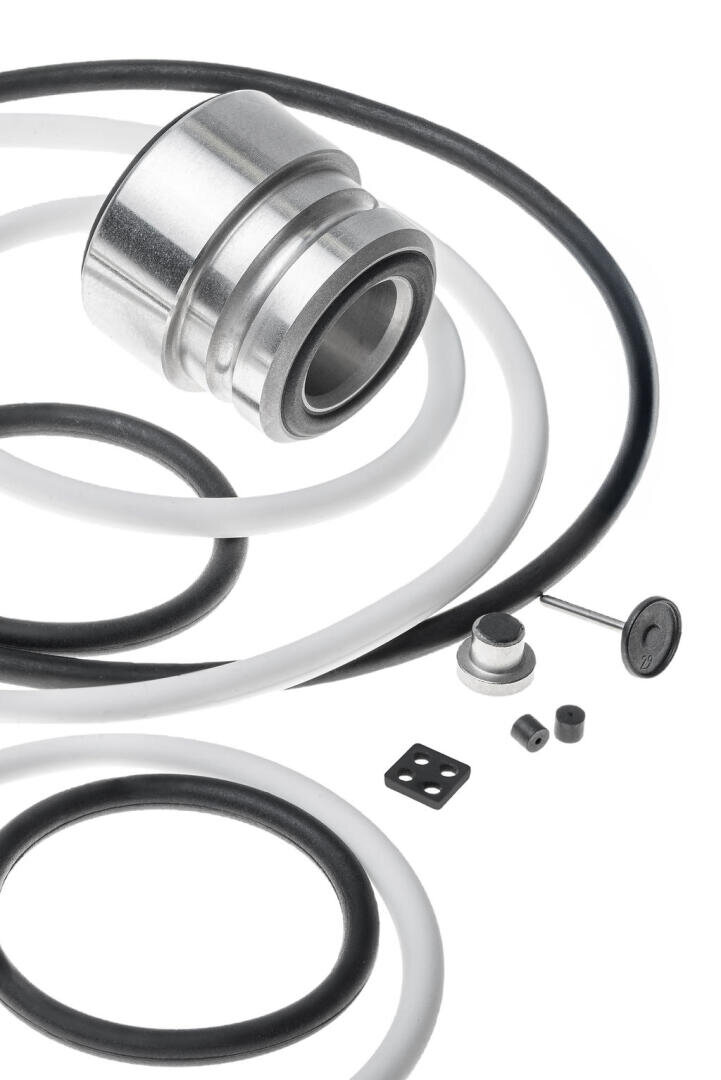 Close-up of various black and white rubber O-rings, a metal cylinder, a valve stem and small seals on a white background, arranged in a visually organized layout.
