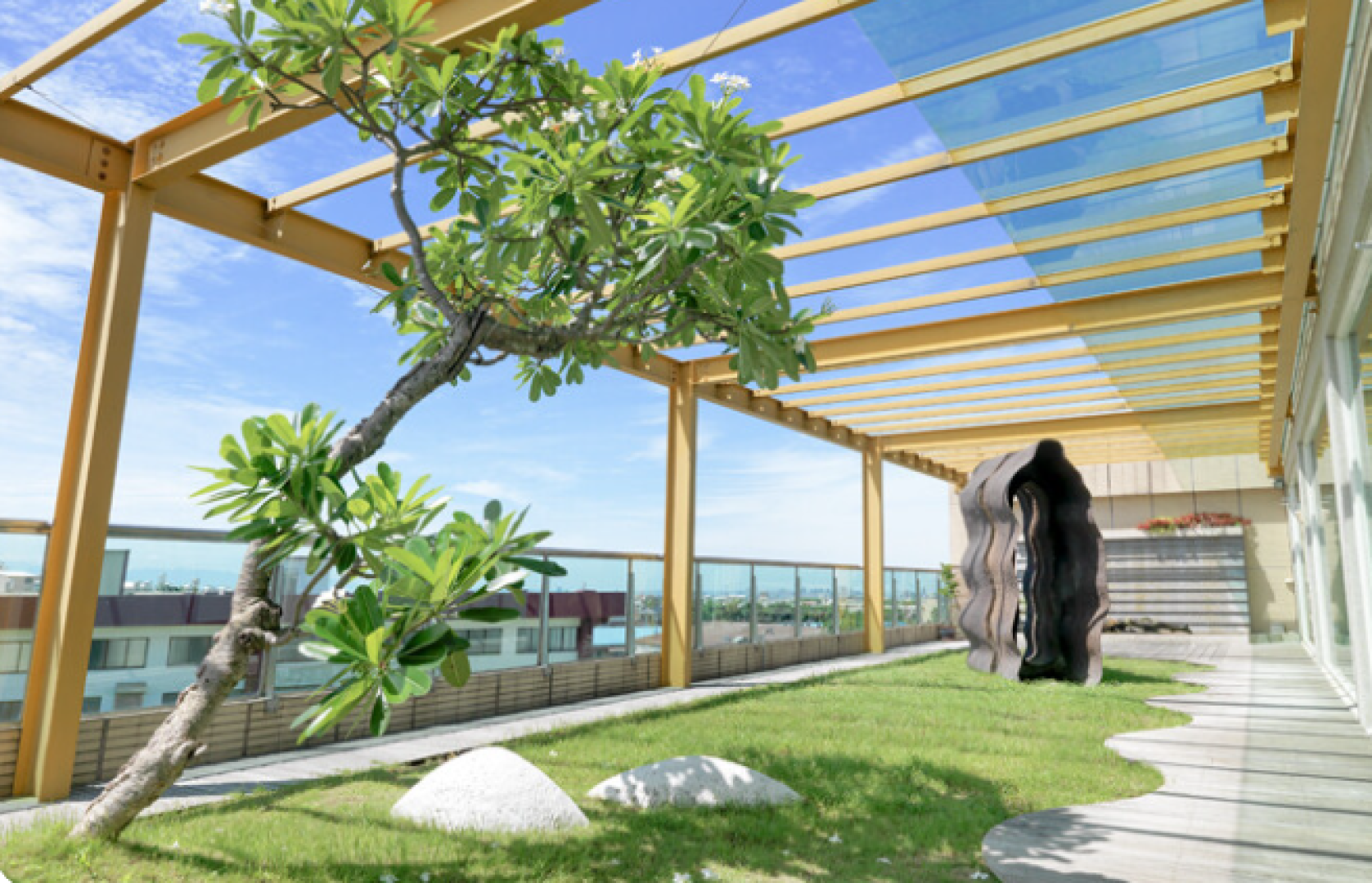 Ein Dachgarten mit grünem Gras, einem kleinen schiefen Baum, weißen Steinen, modernen wellenförmigen Skulpturen und einer gelben Pergola unter blauem Himmel. Glasgeländer säumen die Ränder und geben den Blick auf die umliegenden Gebäude frei.