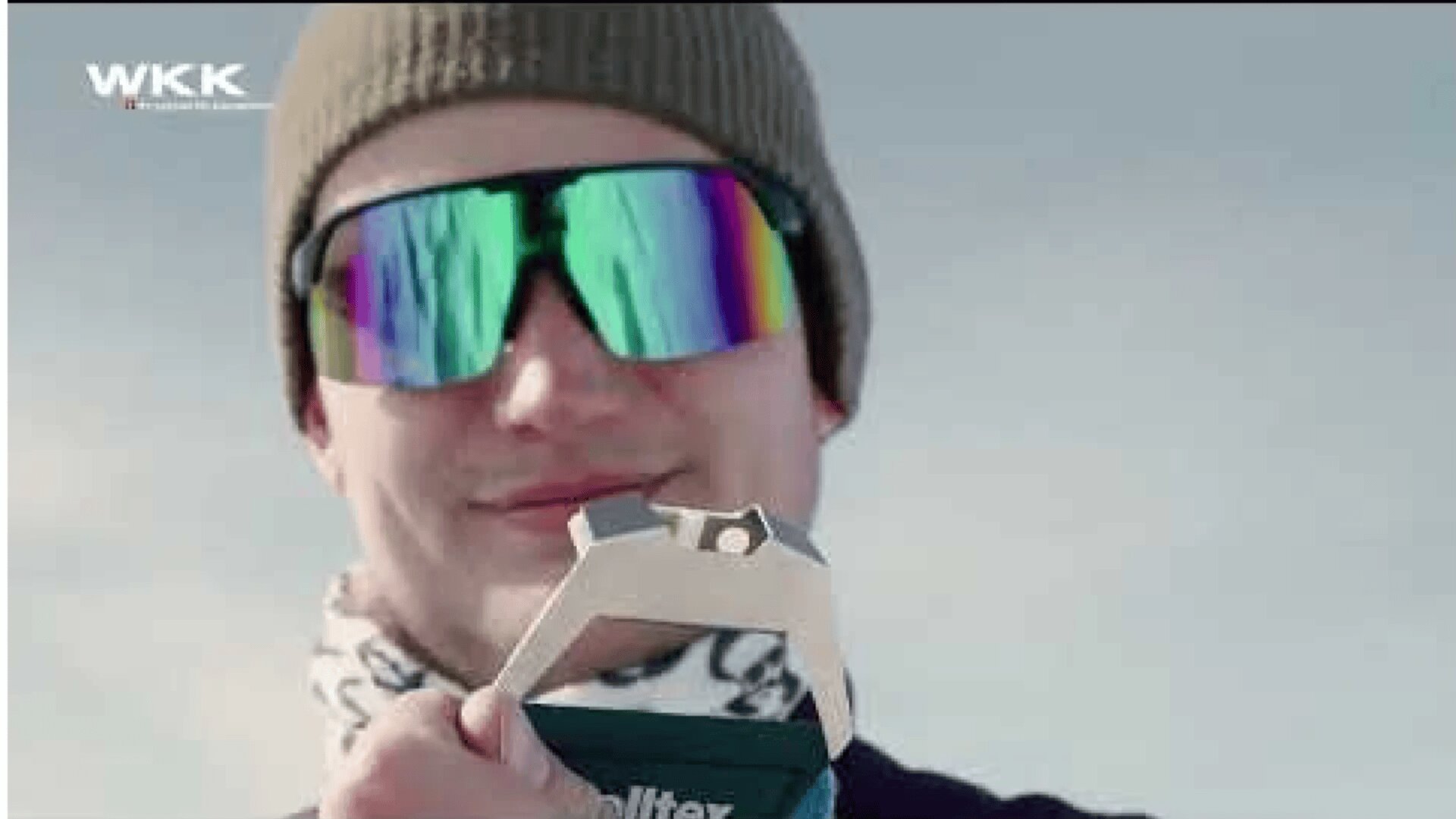 Eine Person mit Beanie-Mütze, verspiegelter Sonnenbrille und gemustertem Schal hält im Freien ein Metallwerkzeug hoch, in der Ecke ist das WKK-Logo zu sehen.