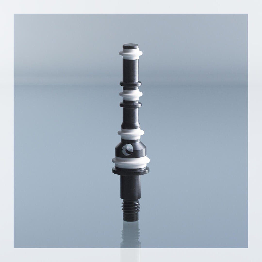 A black and white cylindrical mechanical component with multiple rings and sections stands upright on a reflective surface against a light blue background.